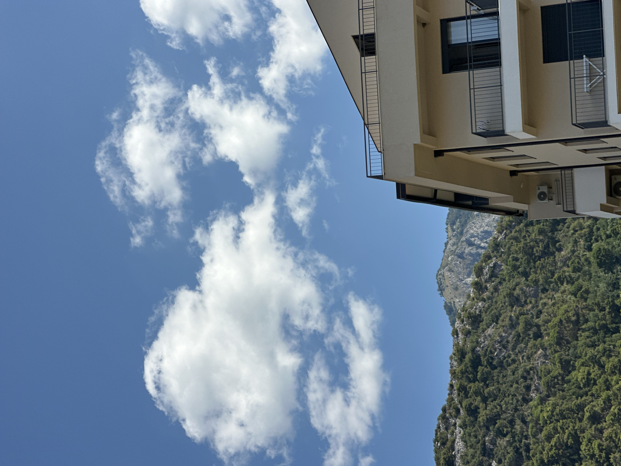 In Budva, Montenegro, the image captures a portion of a modern apartment building in the foreground, characterized by its light beige facade and black railings on its balconies. The building features multiple floors with dark-framed windows and sliding doors leading to the balconies. An air conditioning unit is visible mounted on the side of the building.
In the midground, a densely forested mountainside dominates, with various shades of green indicating a mix of trees and shrubbery. The terrain appears steep and rugged, with visible rocky outcrops interspersed with the foliage.
The background reveals the peaks of the mountains, with some areas showing exposed rock formations, suggesting a dramatic and natural landscape surrounding the urban development. The sky above is a vibrant blue, punctuated by scattered white cumulus clouds, indicating fair weather. The scene is illuminated by natural sunlight, casting subtle shadows on the building and highlighting the texture of the mountainside.