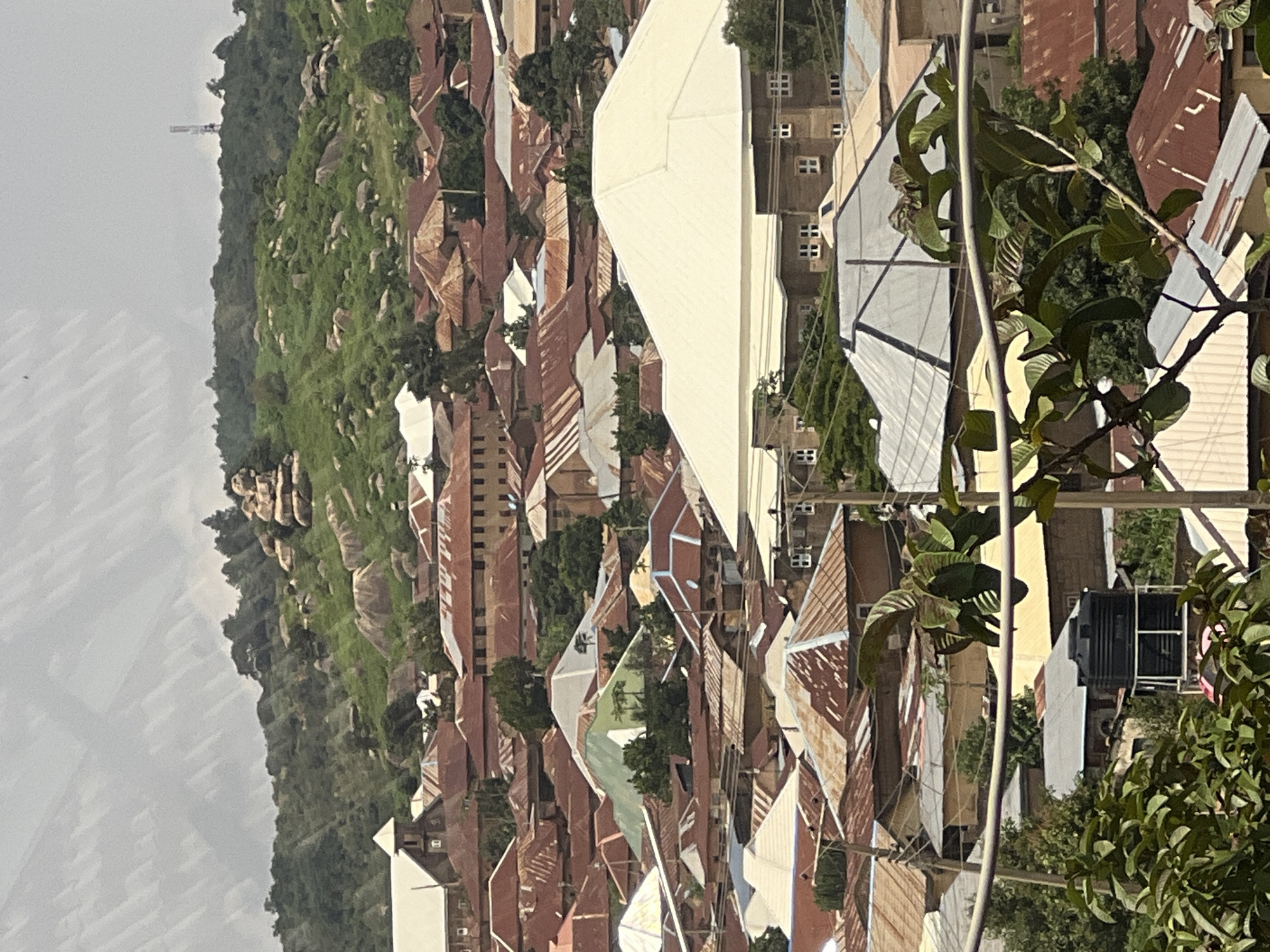This is an aerial, eye-level shot of a densely populated urban area in the city of Jos, Nigeria, characterized by a sprawling collection of buildings with predominantly corrugated metal roofs. The scene is set against a backdrop of lush green, rocky hills, with a telecommunications tower visible on the highest peak. The buildings are mostly low-rise, with a mix of residential and commercial structures. Many of the roofs show signs of age and weathering, with patches of rust and varying shades of orange and brown. Some buildings feature whitewashed walls, while others have a more muted, earthy tone. In the foreground, the branches of a leafy tree extend into the frame, partially obscuring some of the buildings. A large black water tank on a metal stand is visible among the rooftops. Power lines crisscross the scene, adding to the urban infrastructure. The overall impression is one of a vibrant, lived-in community nestled within a dramatic natural landscape.