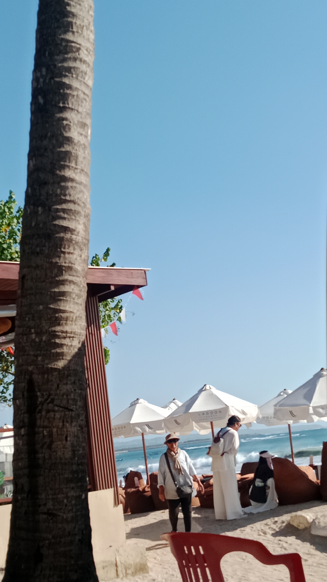 In Kuta, Indonesia, on a sunny day, a group of people are seen on a sandy beach. The scene is dominated by a large palm tree trunk in the foreground, to the left of the image. In the middle ground, several white beach umbrellas are set up, casting shadows on the sand. Behind the umbrellas, the ocean is visible with waves breaking on the shore.
There are at least three individuals in the frame. One person, likely a tourist, stands near the umbrellas wearing a hat, a light-colored long-sleeved shirt, and dark pants, with a crossbody bag. Another individual, dressed in white, stands with their back to the camera, wearing a backpack. A third person, wearing a hijab and a traditional garment, is seated on a large cushion in the background. In the foreground, a red plastic chair is partially visible. Red and white pennant flags hang from a wooden structure above and to the left of the umbrellas. The sky is clear and blue.