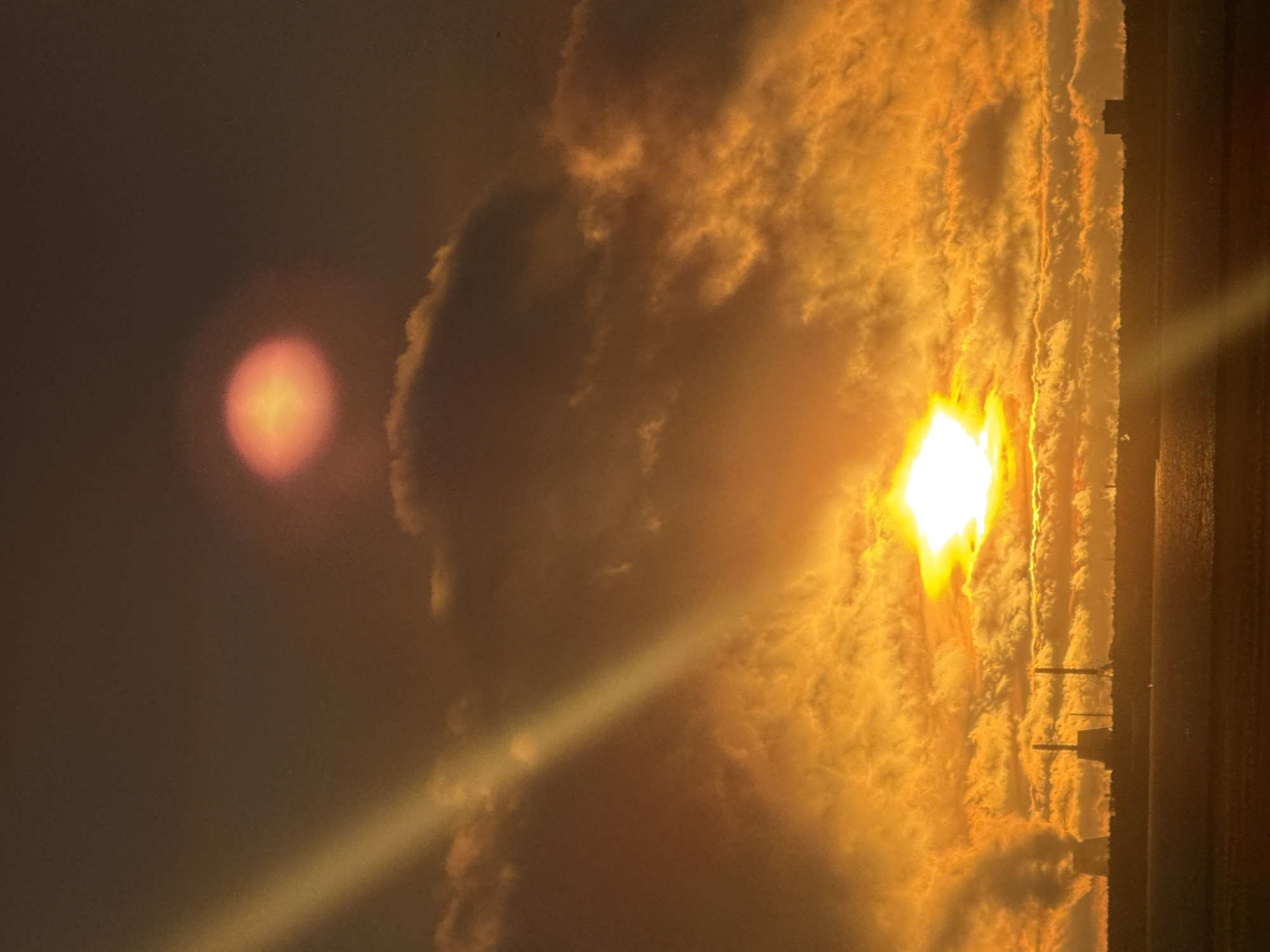 The image captures a dramatic sunset over an industrial landscape, likely in Saint Petersburg, Russia. In the foreground, a dark, flat expanse of land stretches towards a body of water, which reflects the vibrant hues of the sky. Silhouetted against the fiery orange and yellow clouds are industrial structures, including what appear to be cooling towers emitting plumes of steam, and tall, slender chimneys. The sun, a brilliant white orb, is partially obscured by dramatic, textured clouds, casting a strong golden light and creating a visible sunbeam that cuts diagonally across the upper left portion of the frame. A second, smaller, reddish-orange orb, possibly a lens flare or a secondary atmospheric phenomenon, is visible higher in the sky. The overall atmosphere is one of intense natural beauty juxtaposed with industrial activity. There are no people or animals visible in the scene.
