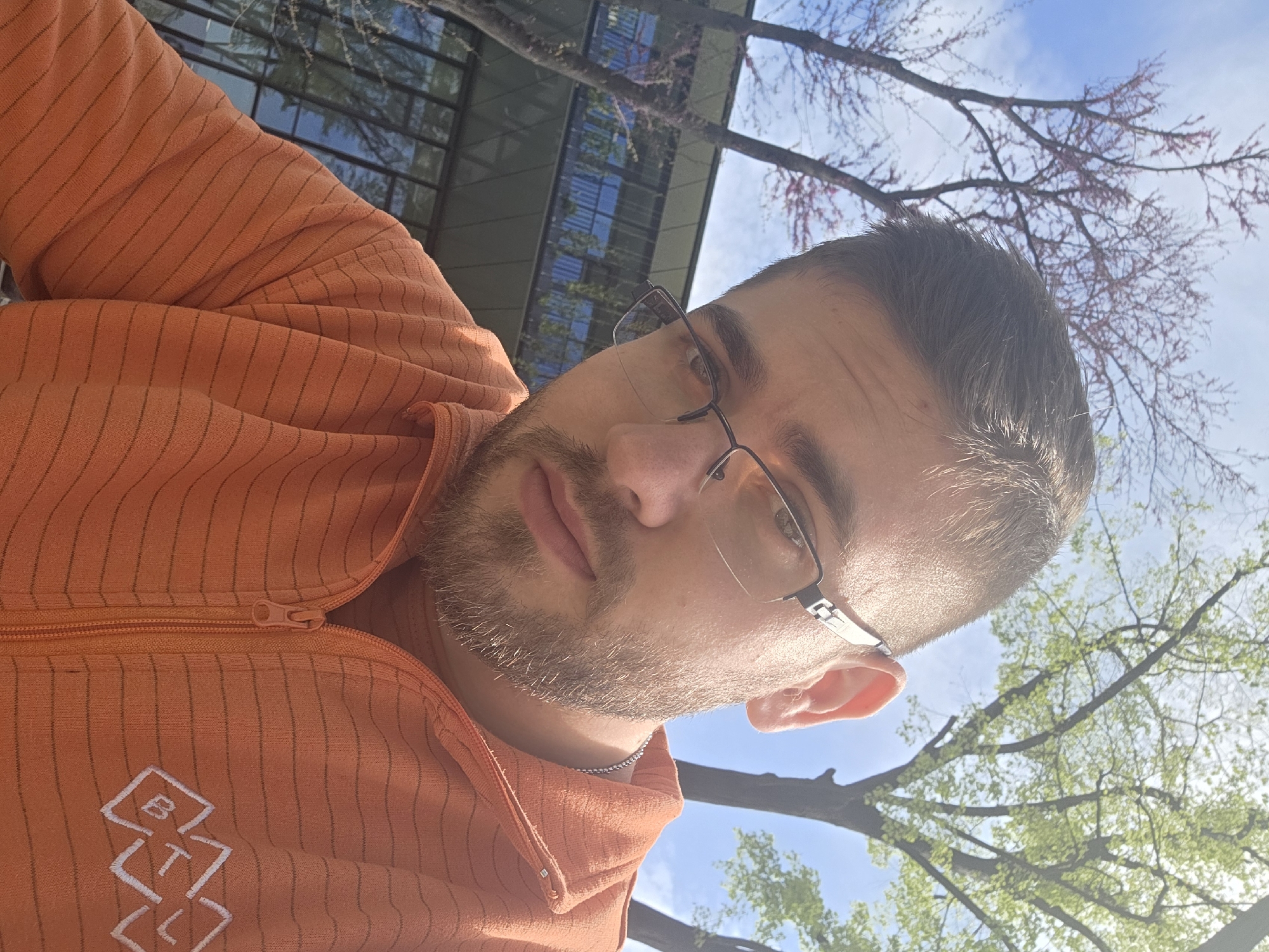 The image captures a bright, low-angle outdoor portrait of a man, likely a selfie, set in what appears to be an urban or campus environment in Plovdiv, Bulgaria. The subject is a young to middle-aged man with short dark hair, a neatly trimmed beard, and glasses, looking directly at the camera with a neutral expression.
He is wearing an orange zippered jacket featuring subtle dark horizontal stripes. On the left side of his chest, a distinctive white embroidered logo is visible, composed of stylized geometric shapes that incorporate the letters 'B', 'T', and 'L'. A silver chain is also visible around his neck.
The background reveals a modern building with reflective glass windows. Surrounding the man and above the building are various trees. Some trees display vibrant green, newly unfurled leaves, while others feature reddish-purple blossoms or are in a budding phase, strongly indicating the spring season. The sky is clear and blue with scattered white clouds, suggesting bright, sunny daytime weather.