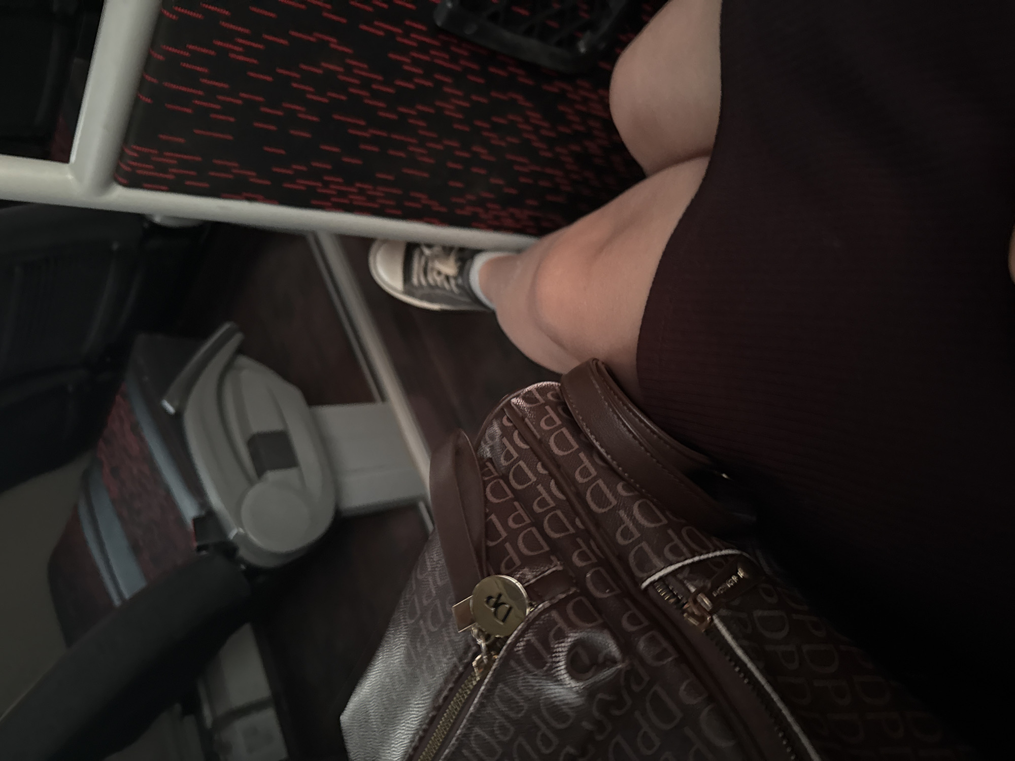 Here is a detailed description of the image:
The image appears to be taken inside a public transportation vehicle, likely a bus or train. A person is seated with a brown patterned handbag or satchel resting on their lap. The person is wearing a dark-colored dress or skirt, and part of their leg is visible, showing a white sock and a grey and black sneaker.
The seat's upholstery has a red and black pattern, and a white frame is visible, suggesting the seat construction. Headphones with a grey cup and black accents are placed on the person's left side, next to the seat. A small portion of a window can be seen behind the headphones.
Based on the prompt, it is implied that the location is in посёлок опытного хозяйства «Ермолино», Russia. However, the image itself doesn't provide any specific visual elements to confirm this location.