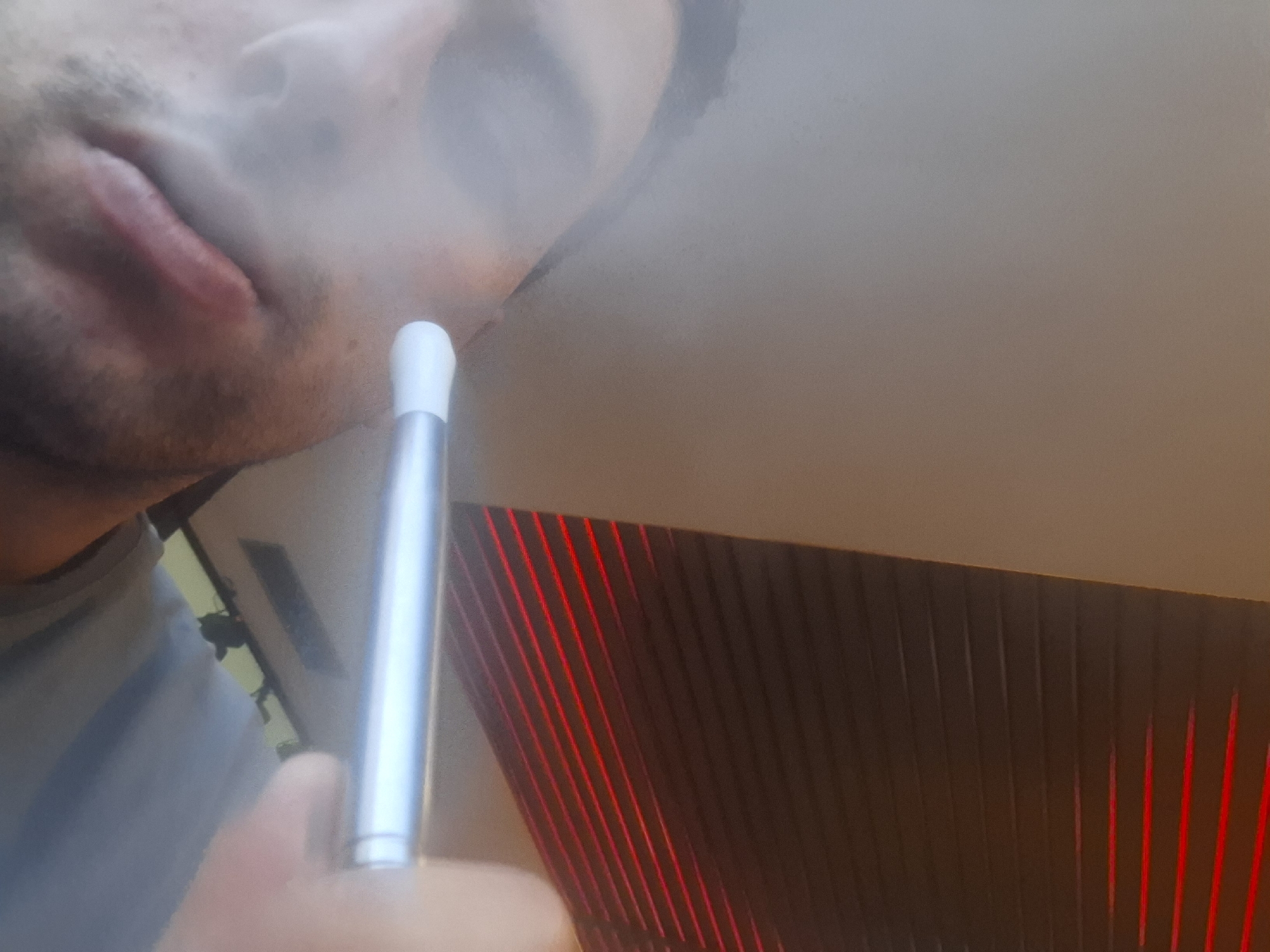 A close-up shot shows a person exhaling vapor from what appears to be a vaping device. The person is cropped, showing only the lower part of their face, including the lips, chin, and part of the nose. Smoke or vapor is visible in the air around their face. The vaping device is silver with a white tip and is held in the person's hand.
The background is out of focus, but it seems to be an interior setting with a wall featuring a design of dark panels and some red light accents. The location in Sofia, Bulgaria is not identifiable from the image.