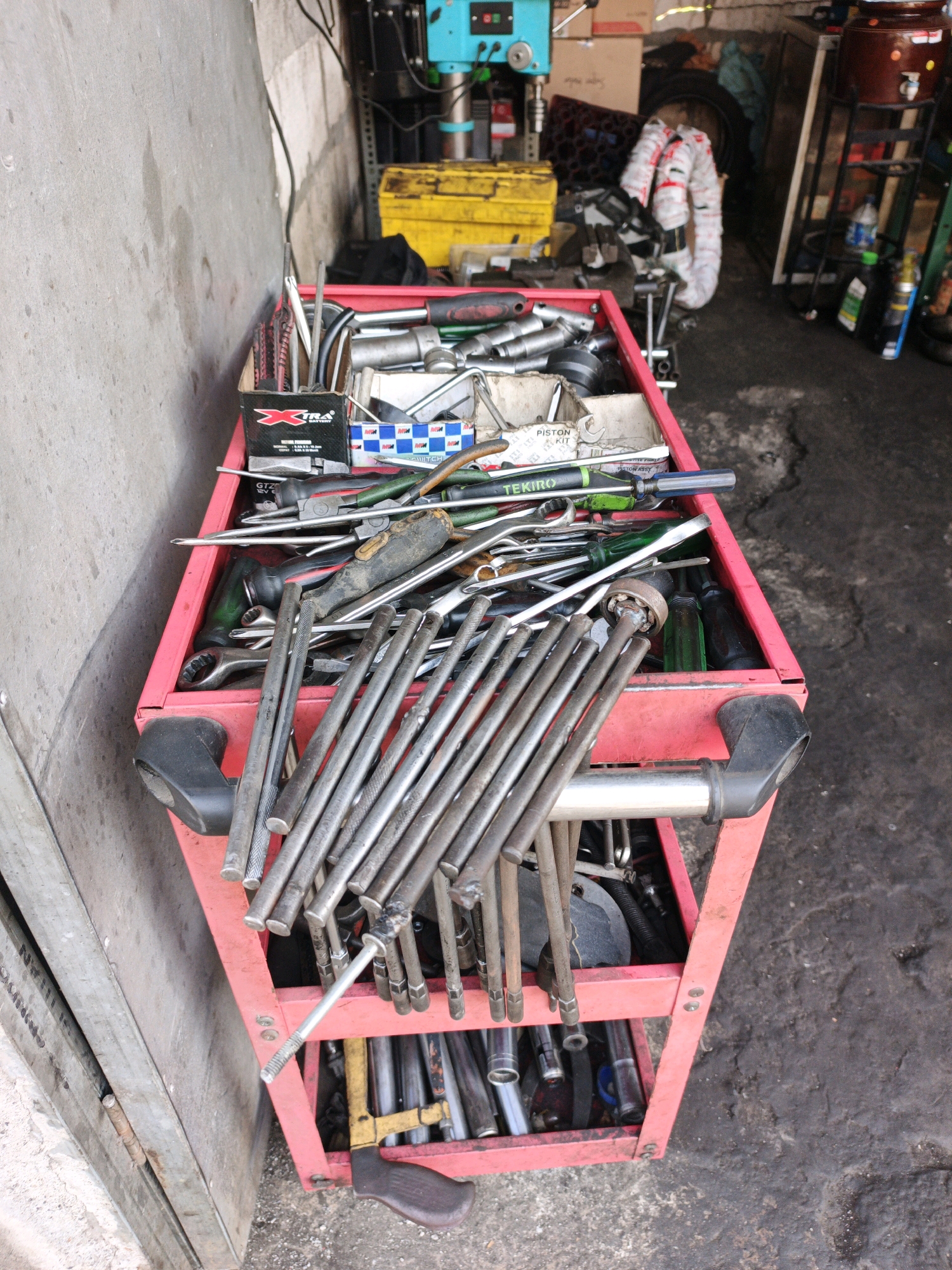 The image depicts a red tool cart filled with various automotive tools, including wrenches, screwdrivers, pliers, and sockets. The cart is situated in what appears to be a workshop or garage, with a concrete floor and a wall on the left side. In the background, there are shelves with boxes, a yellow toolbox, and a blue piece of machinery. To the right of the cart, there's a dispenser for water or other liquids, and some bottles. The scene is consistent with a mechanic's workspace in Boro, Indonesia, an area known for its motor vehicle repair shops. There are no people visible in the image.