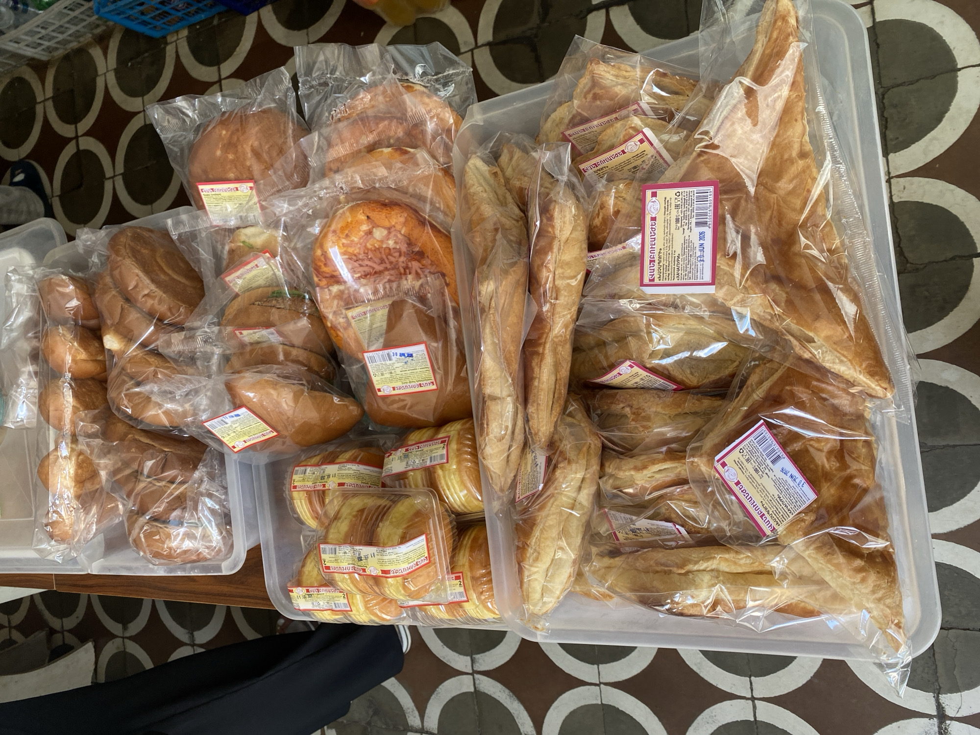 A display of various baked goods, including loaves of bread, croissants, and what appear to be khachapuri, are arranged in plastic trays on a table. The background shows a tiled floor with a repeating circular pattern and a partial view of a person wearing dark clothing on the left. A blue plastic basket is visible on the right side of the image. Some of the items have labels, suggesting they are for sale. The lighting indicates it is daytime, likely outdoors or in a well-lit indoor space. The scene is consistent with a market stall or bakery in Alaverdi, Armenia.