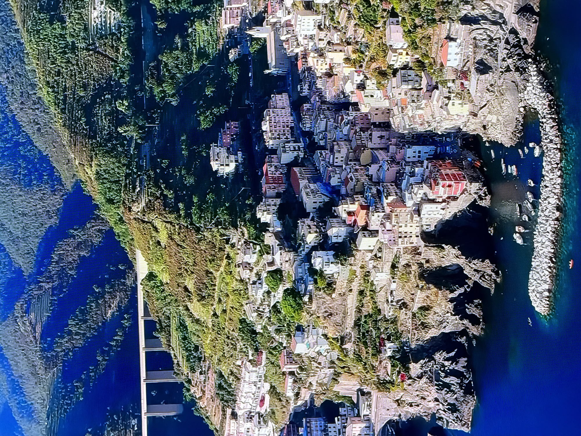 This aerial photograph captures a densely populated coastal village nestled on a steep, terraced mountainside overlooking a deep blue body of water. The buildings are colorful, tightly packed, and built on multiple levels, cascading down towards the water. Many appear to be residential, with small windows and tiled roofs.
Dominating the landscape is the vibrant green of the terraced hillsides, indicating agricultural activity, likely vineyards or olive groves. A modern, multi-span concrete bridge is visible in the upper left quadrant, spanning across a gap in the hills and connecting to the village's periphery. The water below is calm, with several small boats docked or anchored. A breakwater constructed of large stones juts into the water, providing protection to the harbor. The scene is bathed in sunlight, casting shadows that highlight the rugged terrain and the architectural density of the village.
There are no discernible people or specific landmarks that would suggest this location is the city of Chirchik, Uzbekistan.