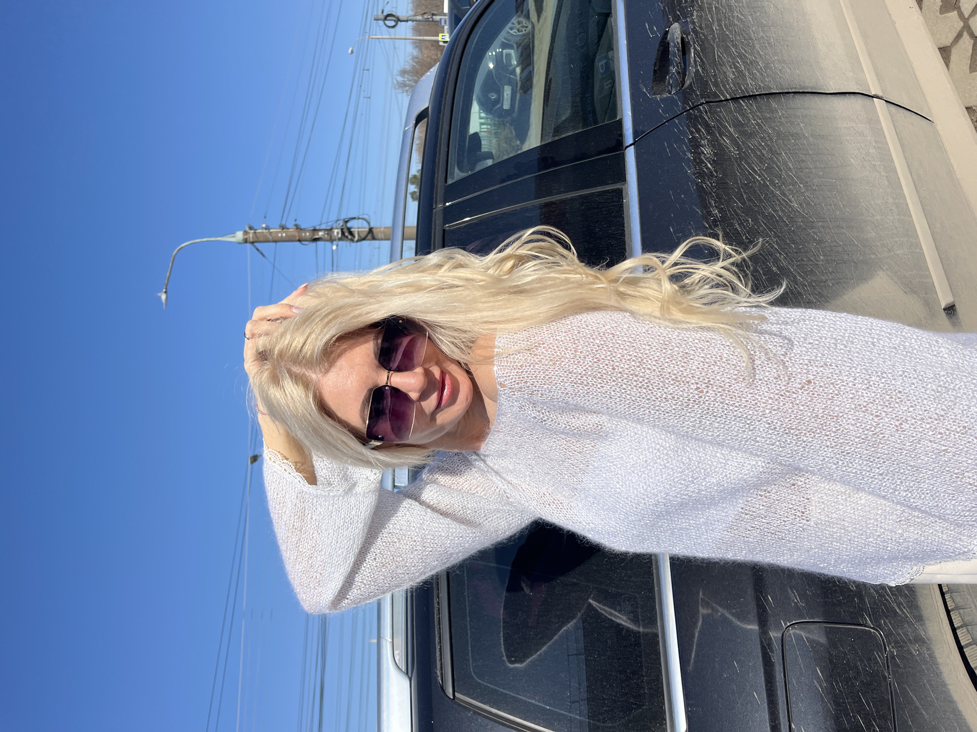 A blond woman in sunglasses and a white sweater stands next to a dirty black SUV under a clear blue sky. She has her hands behind her head. Overhead utility lines and a streetlamp are visible. The location is likely in Nizhny Novgorod, Russia, based on visual cues such as architecture and vehicle types common in the region.