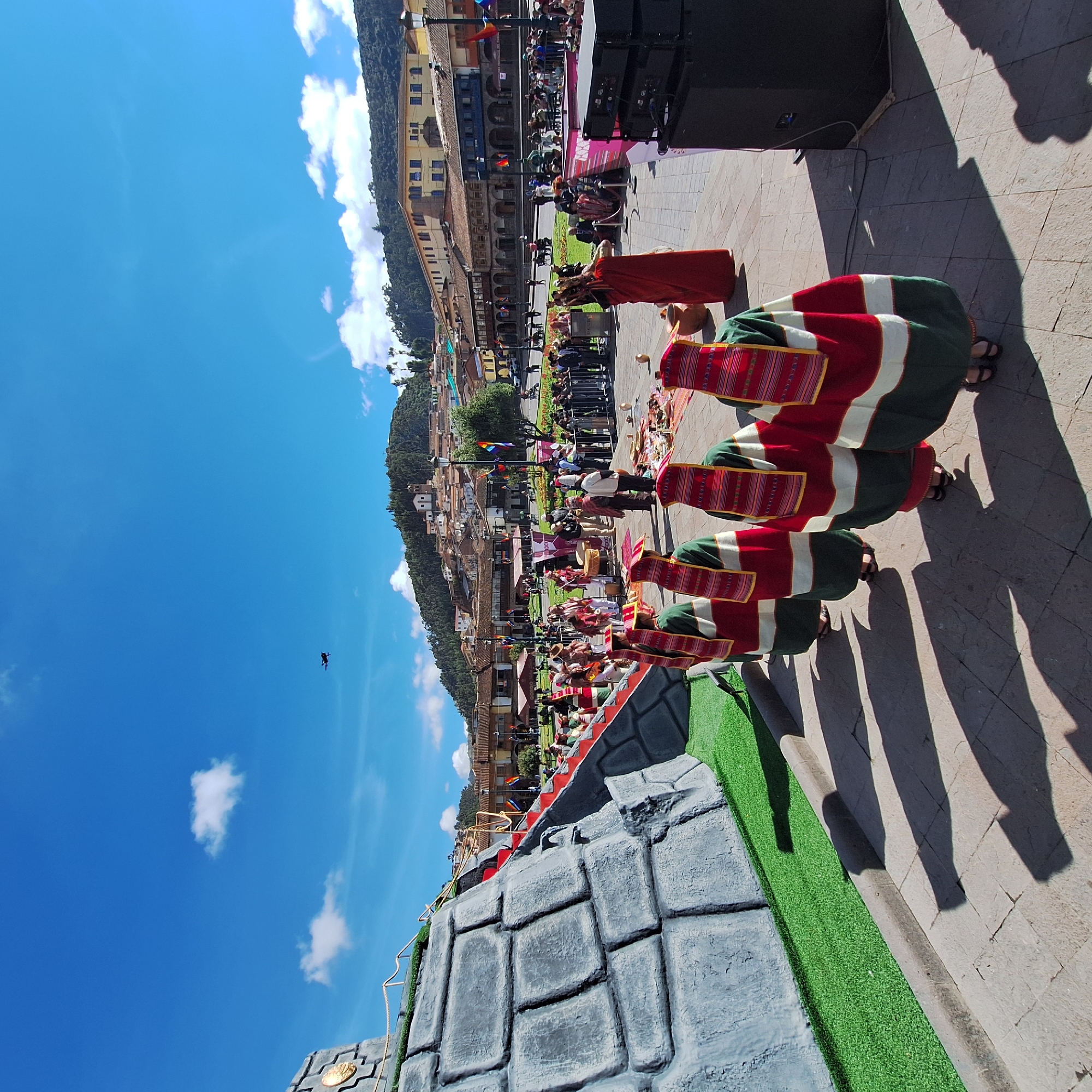 This photograph shows a cultural event in a public square in Cusco, Peru. In the foreground, a faux-stone barricade with synthetic green turf lines the edge of the stage. Three individuals wearing traditional Andean clothing, including red, green, and white striped textiles, stand in a row. Their shadows stretch across the cobblestone pavement, indicating a strong light source. The square is crowded with spectators standing behind a metal barricade, observing the performers. In the background, a building displays the rainbow flag, and more spectators are gathered along the upper floors of the buildings. A wooded hill slopes upward behind the urban landscape, and a clear blue sky with scattered clouds fills the upper portion of the frame. A dark bird is visible mid-flight in the upper left corner.