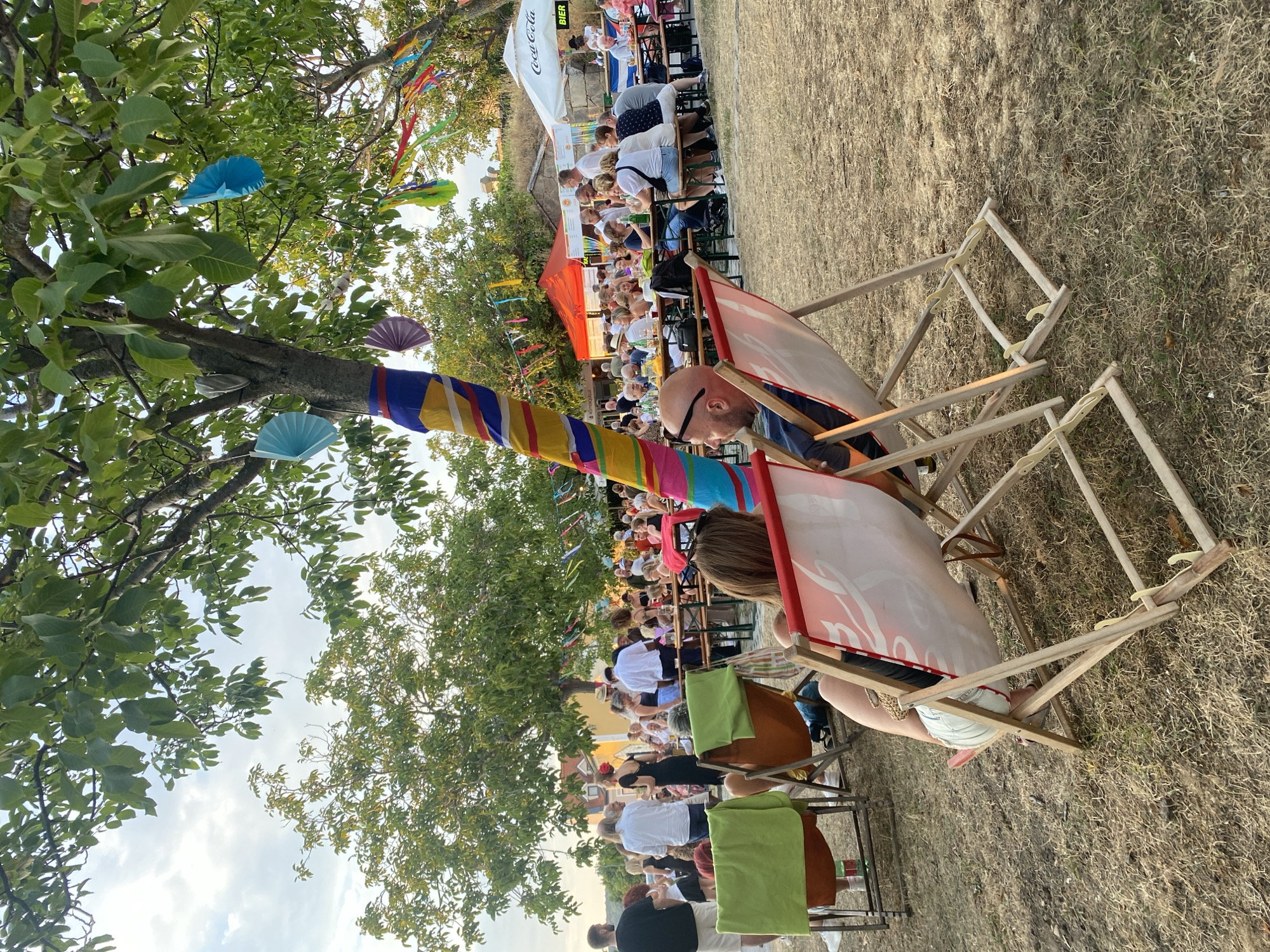 The image depicts an outdoor event in Purbach, Austria, likely a summer festival or gathering, set under a large tree adorned with colorful decorations. In the foreground, two individuals are seated in deck chairs, facing away from the camera, enjoying the festivities. One person, a man with sunglasses, is looking towards the right side of the frame. The other person, a woman, is partially visible, with her legs crossed. Behind them, a lively scene unfolds with numerous people gathered at long tables, engaged in conversation and dining. Tents and a Coca-Cola branded umbrella are visible in the background, suggesting food and beverage services. The overall atmosphere appears relaxed and social, with a backdrop of trees and buildings, indicative of a community event taking place in the historic town of Purbach.