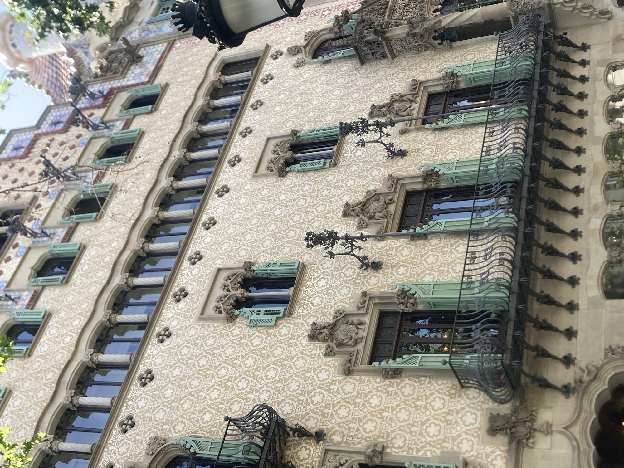 The image shows an exterior view of a highly ornate building, likely a residential or commercial structure. The architecture features intricate detailing, with a patterned facade in a cream and light brown color. Repeating floral or geometric motifs are visible across the surface.
Numerous windows are present, many of which are framed with decorative elements. Some windows have external balconies with elaborate black metal railings, featuring a wavy design. Small, light green awnings or shutters are visible above and below some of the windows, adding a touch of color to the facade.
Decorative sculptures and architectural embellishments are integrated into the building's design, particularly around the windows and cornices. These ornate features suggest a historic or art nouveau style. In the upper left corner, some greenery from trees is visible, along with a glimpse of a blue sky, indicating an outdoor scene, likely during daylight. The overall impression is one of a visually rich and architecturally significant building. No people are visible in the image. There is no visible text. The scene is likely set in Barcelona, Spain, given the architectural style which is characteristic of the city.