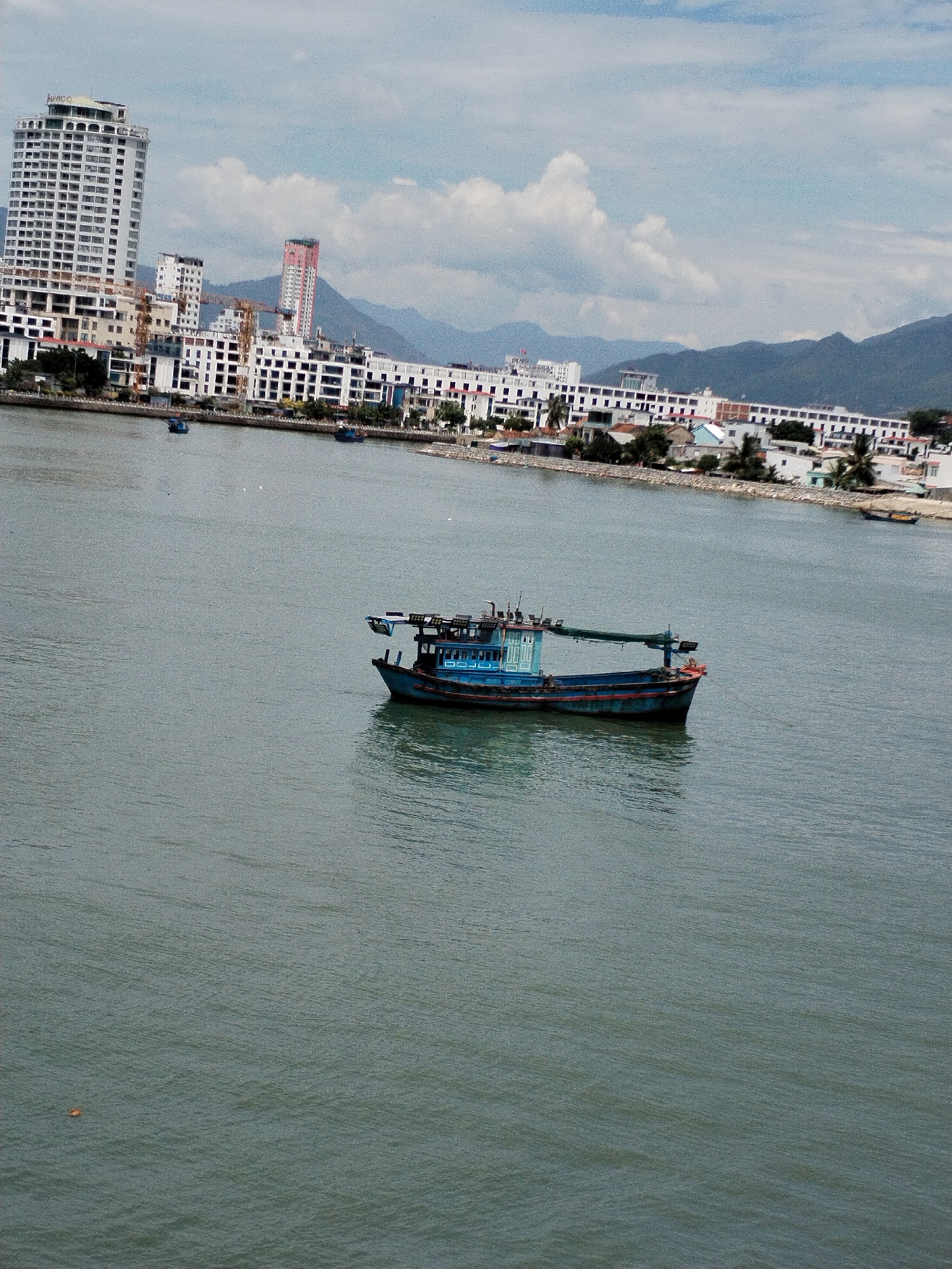 The image depicts a scene in Nha Trang, Vietnam, featuring a body of water with a blue fishing boat in the foreground. In the background, a city skyline is visible, characterized by tall buildings, including a prominent white high-rise. Mountains form the backdrop to the city, and the sky is partly cloudy. The water appears calm, with the boat casting a reflection. The overall atmosphere suggests a daytime view of a coastal urban environment.