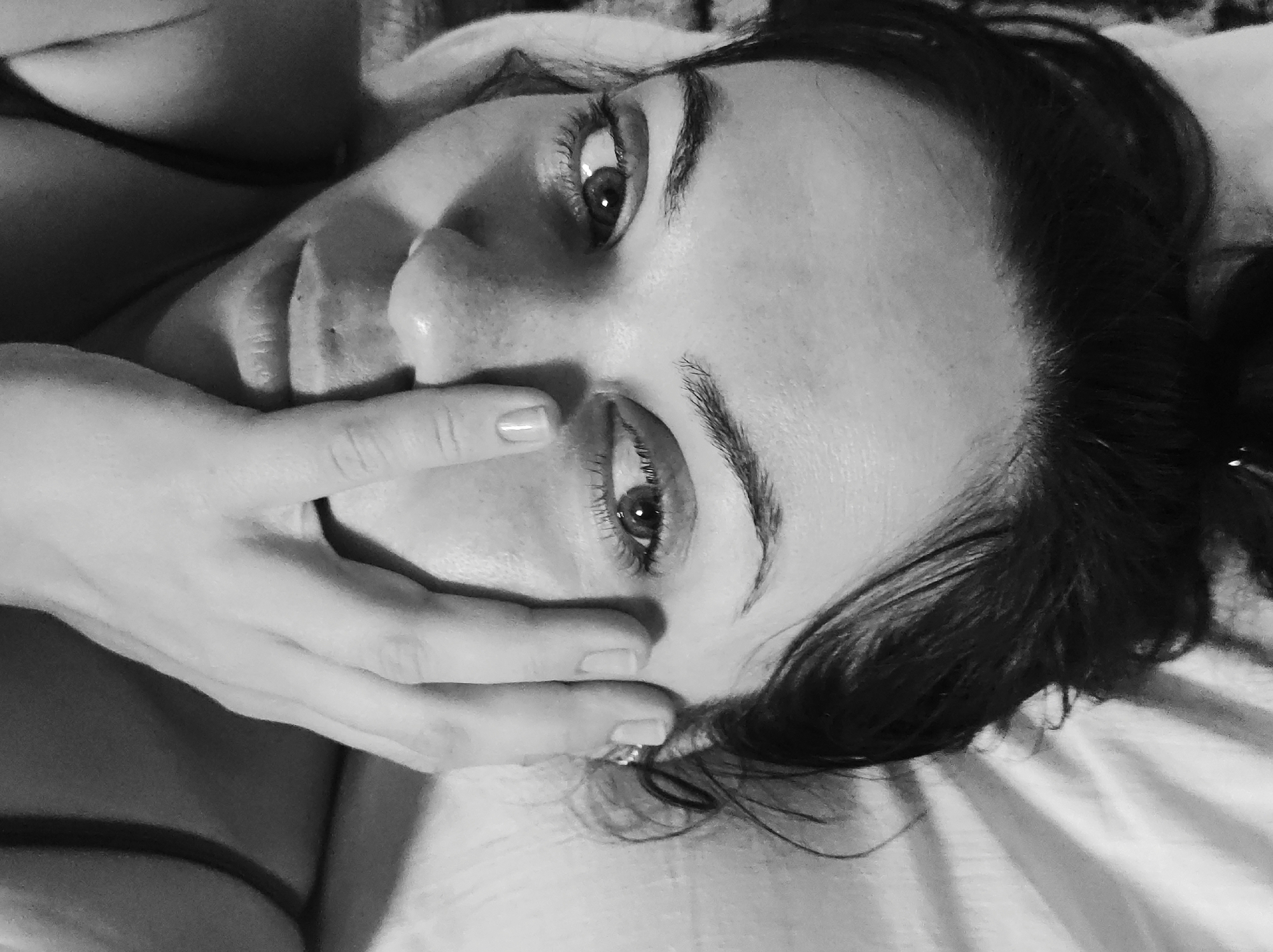 The black and white image features a female subject lying down, with her head oriented towards the right. Her eyes are open and directed upwards and to the viewer's left, with light reflections visible in her pupils. Dark hair frames her face, and distinct eyebrows are present. Her left hand is positioned across the lower part of her face, covering her mouth and chin. The hand displays manicured nails and a ring on the ring finger. A dark strap from clothing is visible on her upper left chest. The subject rests on what appears to be a bed with light-colored, creased sheets. A darker surface is visible behind her head. The lighting is soft, creating moderate shadows and highlights. No visual elements within the image content identify its location as Фоки, Russia.