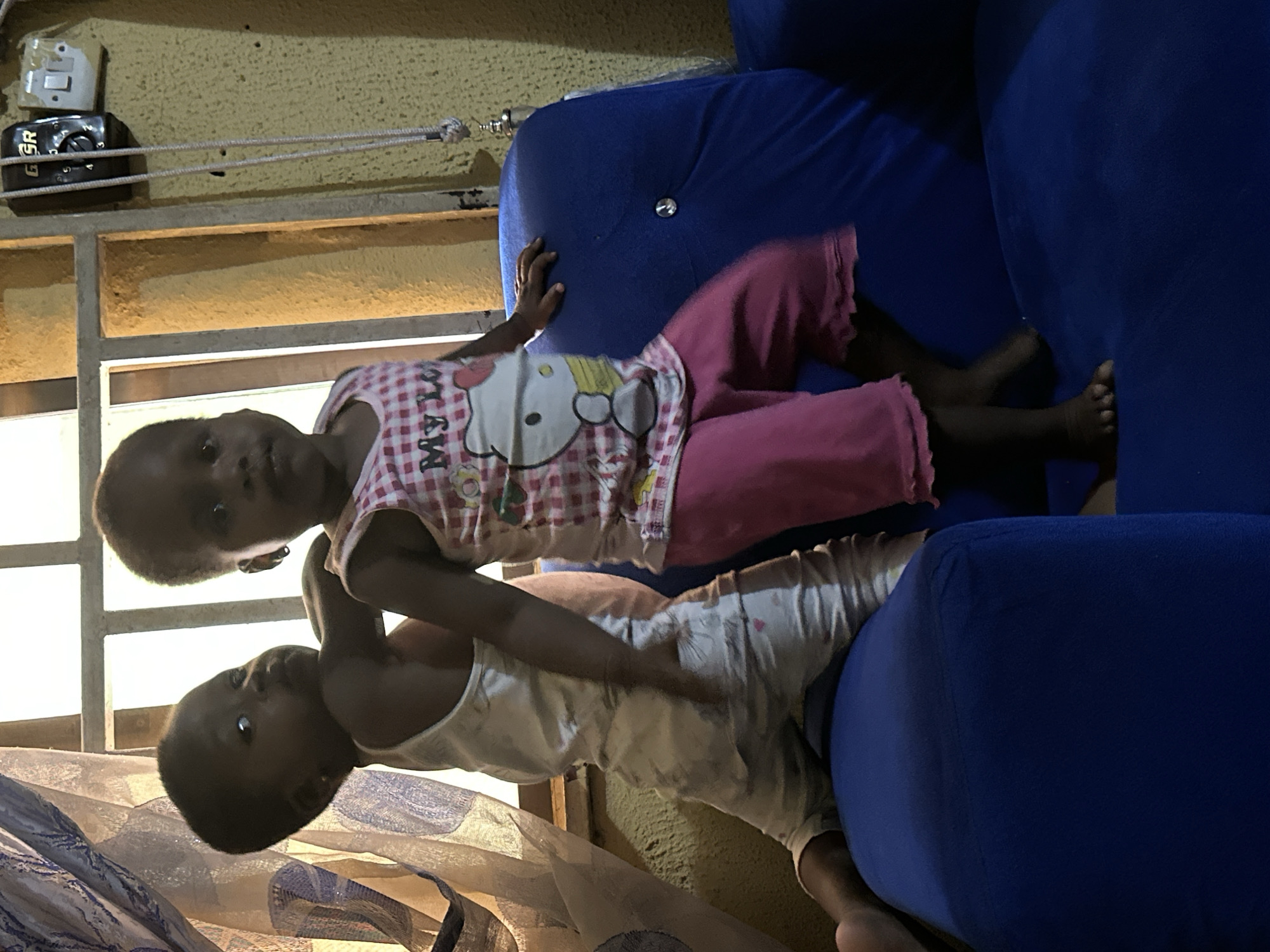 Two young Black children are on a blue couch in a room in Narayi, Nigeria. The child on the left, wearing a white tank top and white pants with pink hearts, is leaning forward. The child on the right, wearing a pink and white plaid shirt with a Hello Kitty graphic and pink pants, is standing. The room has a window with a patterned curtain on the left and a yellow textured wall on the right, where an electrical outlet and a dark-colored device are mounted. The children are illuminated by natural light coming from the window.