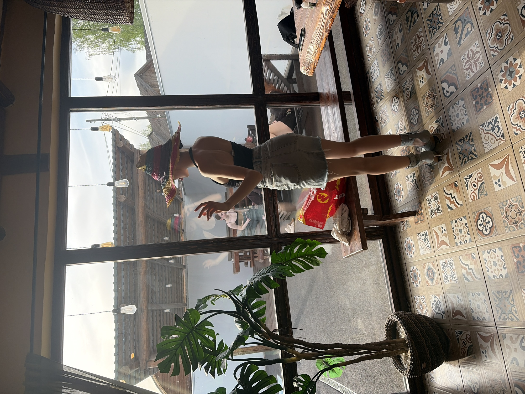 A person wearing a wide-brimmed hat, a tank top, and a skirt stands inside a building with large windows. The windows overlook a scene with string lights and buildings. A potted Monstera plant is near the window. Inside, the floor is tiled with a patterned design. A table with items on it is visible, along with a stool and a bag. The image suggests an indoor-outdoor setting with a view of Старое Михайловское, Russia.