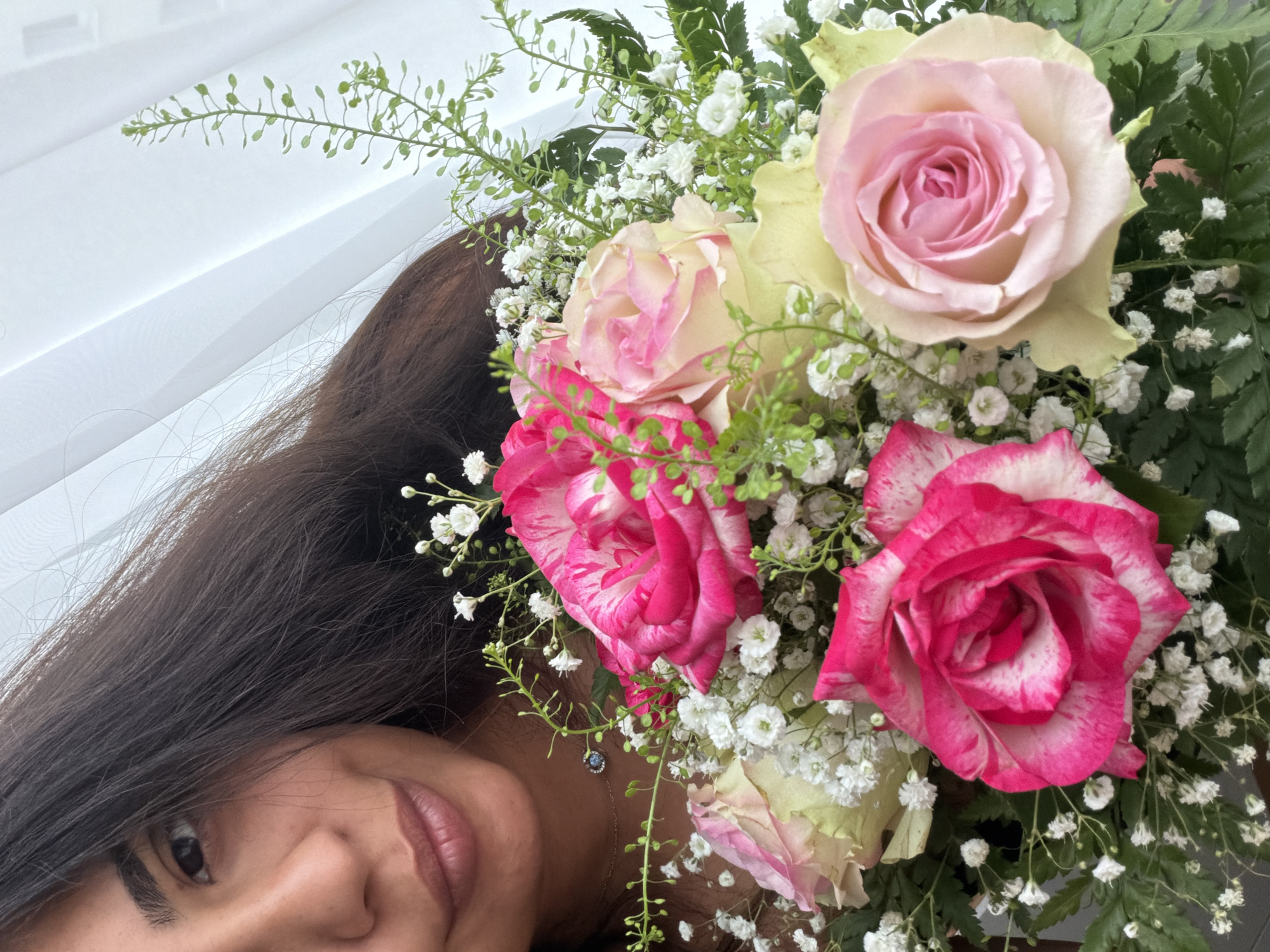 A woman is holding a bouquet of roses and baby's breath. She is looking at the camera with a slight smile. The bouquet is in the foreground, and the woman is in the background. The roses are pink and white, and the baby's breath is white. The woman has long, dark hair, and she is wearing a necklace with a blue pendant. The background is a white curtain. The image does not contain any information that allows for the identification of a specific location in Duisburg, Germany.