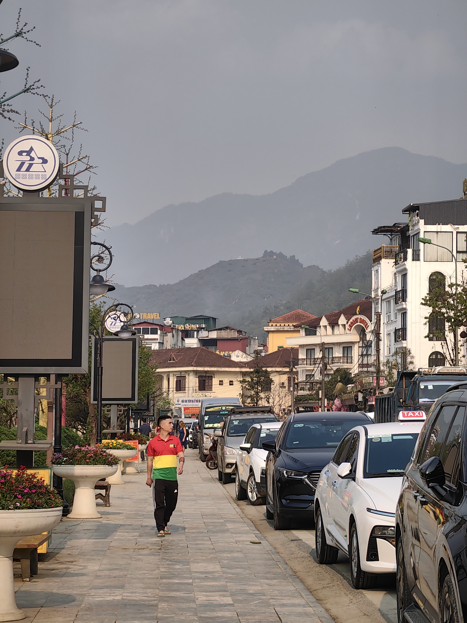 The image depicts a daytime street scene in Sa Pa, Vietnam. A paved pedestrian sidewalk, adorned with large white planters containing colorful flowering plants and wooden benches, runs along the left side. On the far left, a tall grey sign structure features a prominent white circular logo with blue abstract shapes and a large, blank rectangular display panel below it. Decorative metallic structures resembling bare trees with lights are integrated into the signage.
A lone East Asian male, wearing a red, yellow, and green horizontally striped polo shirt, black track pants, and light-colored flip-flops, walks from left to right on the sidewalk, looking forward. His hair is short and dark.
To the right of the sidewalk, a line of parked cars extends into the distance. Visible vehicles include a black SUV, a white sedan, and parts of other vehicles, including one with a "TAXI" sign on its roof.
Beyond the parked cars, multi-story buildings line the street. Several appear to be hotels, identified by signs such as "LEGACY HOTEL" and "TRAVEL." These buildings exhibit varied architectural styles, including white facades with black balconies, arched windows, and tiled roofs. Ornate black streetlights with decorative scrolls and globes illuminate the street.
In the background, large, hazy mountains rise against a light grey sky, characteristic of the mountainous terrain surrounding Sa Pa. The overall lighting suggests either an overcast day or late afternoon.