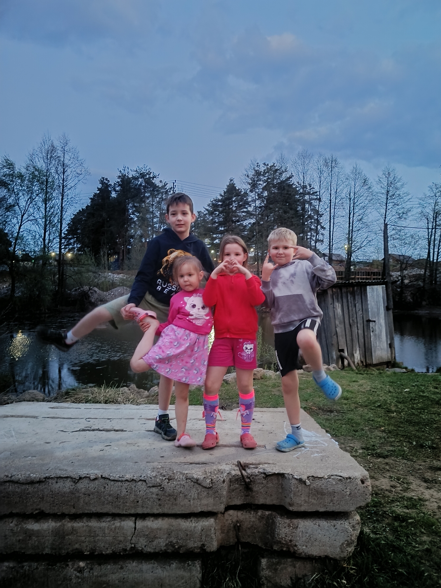 Four children are on a concrete platform. An older boy in a dark hoodie holds a smaller girl in a pink dress with a cat on it. Another girl in a red sweatshirt and shorts makes a heart shape with her hands. A boy in a gray sweatshirt and black shorts strikes a pose with one leg raised. In the background are trees, a body of water, and a small shed. The sky is overcast. This image was likely taken in the city of Chaikovsky, Russia.
