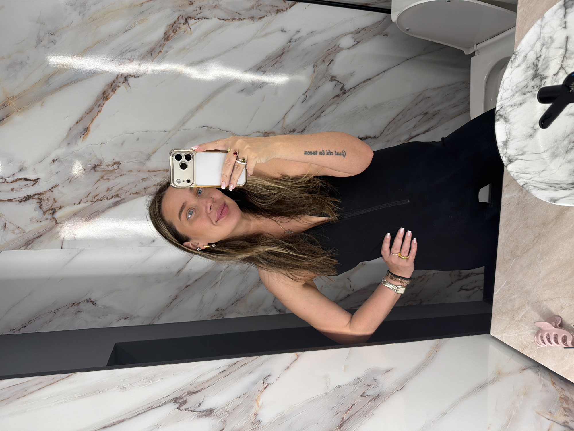 A woman is taking a selfie in a bathroom. The bathroom has marble walls and countertops. There is a toilet and sink in the background. The woman is wearing a black dress. She has a tattoo on her arm. She is holding a phone in her hand. The location is unknown, the city of Moscow, Russia is not discernible.