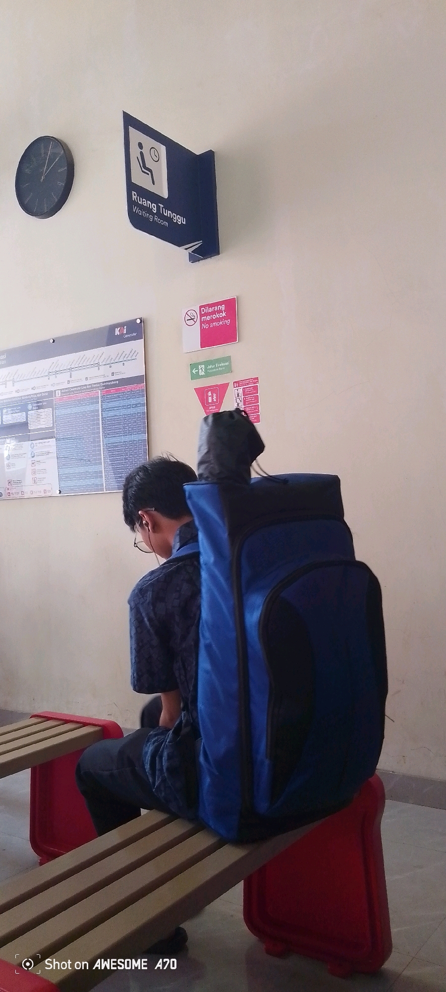 An exterior shot shows a young person sitting on a bench in what appears to be a waiting area at a train station. The individual is facing away from the camera, wearing a blue patterned shirt and dark pants, and has a large blue backpack strapped to their back. To the left of the person, a clock hangs on the wall, and above that, a blue sign indicates "Ruang Tunggu / Waiting Room." Further to the right, a pink sign reads "Dilarang merokok / No smoking," and below it, a green arrow points to "Jalur Evakuasi / Evacuation Route." A large timetable or route map is mounted on the wall behind the person. The environment appears to be clean and well-lit, with a tiled floor and beige walls. This scene is located in Gadobangkong, Indonesia.