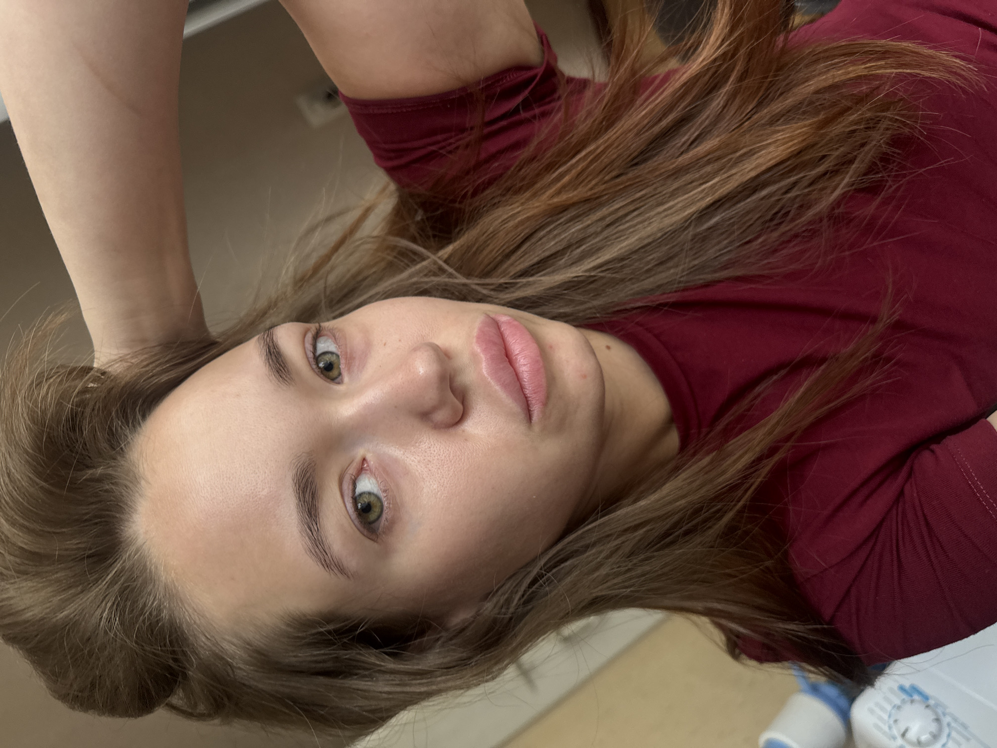 A young woman with long, brown hair pulled back on top of her head, is shown in a close-up, selfie-style shot. She is wearing a dark red t-shirt and her right arm is raised, with her hand touching the back of her head. The background appears to be an indoor setting, with a plain wall and a hint of what might be an appliance on the left side of the frame. The lighting is soft and natural. The image does not contain any identifiable landmarks or elements that would allow for its specific location in Chaikovsky, Russia.