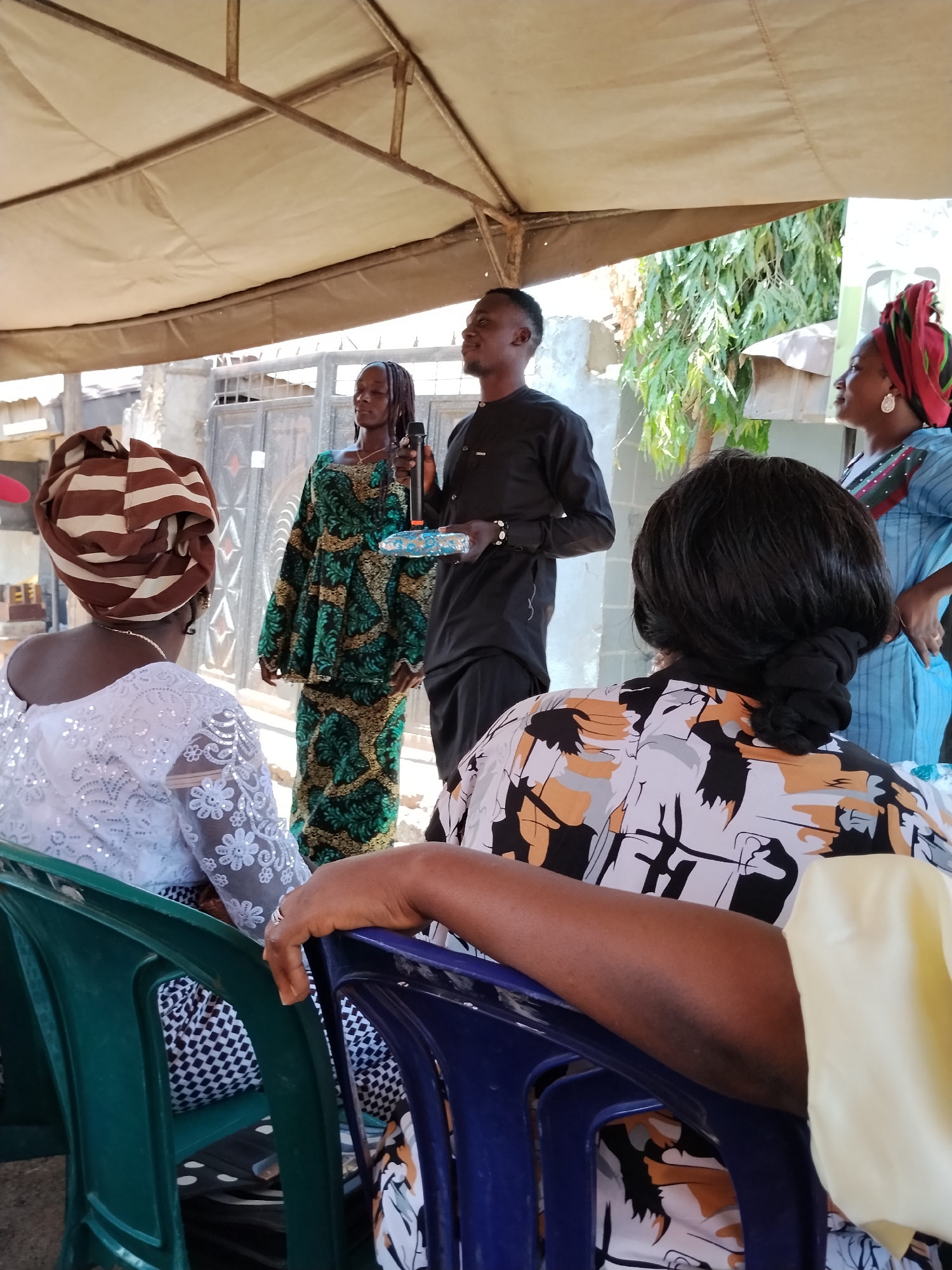 The image shows a gathering of people in Sabon Tasha, Nigeria. A man in a black shirt and pants is holding a microphone and appears to be speaking to the group. A woman in a green and gold outfit stands next to him. Another woman in a blue dress and a red headwrap stands to the side. Several people are seated in plastic chairs facing the speakers. One woman in a white lace top with a brown and white headwrap is visible from the back, as is another woman in a patterned top. The scene is set under a tent-like structure, and a building can be seen in the background. There are green trees in the background.