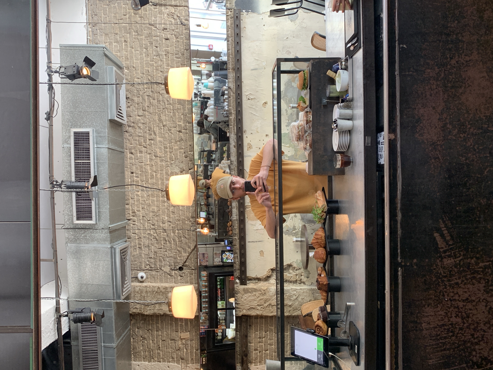 A person is photographing their reflection in the window of a bakery/coffee shop. The reflection shows a street with brick buildings in Tel-Aviv, Israel. The bakery window displays pastries. Inside, one can see pastries and coffee cups. The person in the reflection wears a tan shirt and a baseball cap.