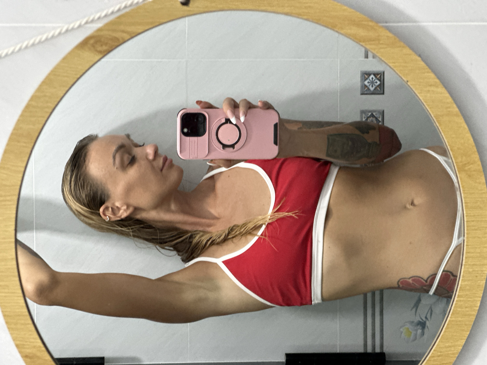 A woman with wet blonde hair pulled back into a braid is taking a mirror selfie in a bathroom. She is wearing a red sports bra with white trim and white underwear. Her right arm is raised above her head, and she is holding a pink phone in her right hand. The bathroom has white tiled walls with a few decorative tiles. A framed mirror with a wooden frame is prominently displayed. The reflection shows the woman's profile, tattoos on her arm and hip, and the phone she is using. The image quality is somewhat blurry due to the mirror reflection. The setting appears to be a residential bathroom, possibly in a hotel or guesthouse, given the tiled walls and decorative elements. There is no clear indication of the city of Mũi Né, Vietnam, or any specific details that would place the image there.