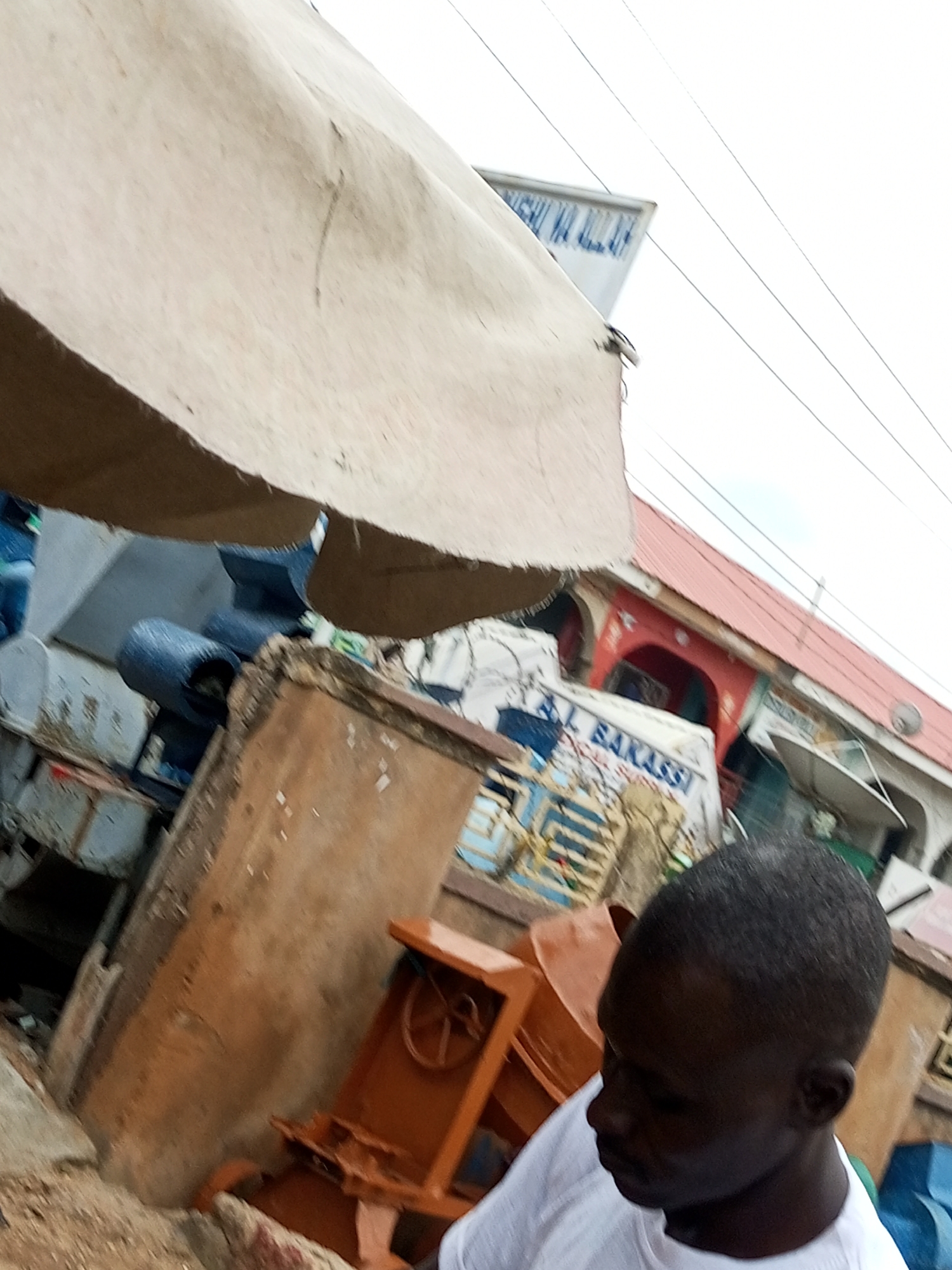 A medium shot, eye-level, outdoor photograph shows a man in a white t-shirt facing right. He has dark skin and short, dark hair. He is in the foreground, slightly blurred, looking down and to the right. Behind him, there are several objects. A tan-colored awning is draped overhead, obscuring the sky in the upper left of the frame. To the right of the awning, power lines are visible against a hazy white sky. In the middle ground, a brown, cement-like structure is visible, possibly a wall or the side of a building. To the left of this structure, a large, dark blue, cylindrical object is partially visible, likely a tank or container. A portion of an orange metal object, possibly a piece of machinery or equipment, is in the lower right quadrant of the frame, also slightly blurred. Further in the background, there are buildings with red roofs and arched doorways. A sign with blue and white lettering, partially obscured, is visible on a white surface. There are also some blue and white striped patterns visible on a structure to the left of the frame. The overall scene appears to be in an outdoor market or industrial area.