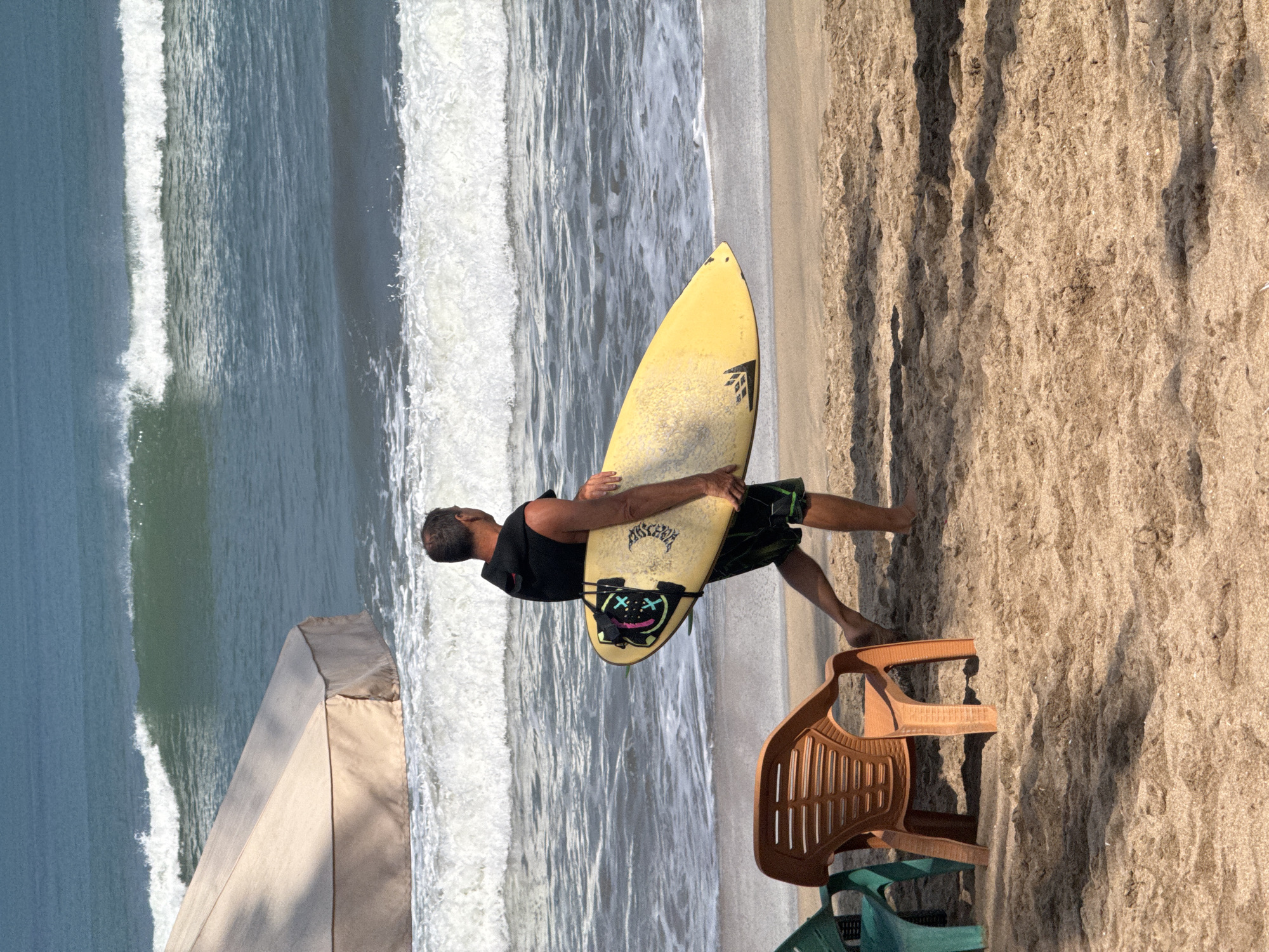 A surfer walks along the sandy beach in Kuta, Indonesia, carrying a yellow surfboard. The surfer, a man with short brown hair, wears a black sleeveless hoodie and patterned shorts. The surfboard has a black strap around it and a design on the bottom featuring two cartoonish faces with "X" eyes. In the background, the ocean features moderate waves and white foam. A tan beach umbrella is partially visible on the left, and two plastic chairs, one orange and one green, sit on the sand in the foreground. The sand is textured with shadows cast by the objects.