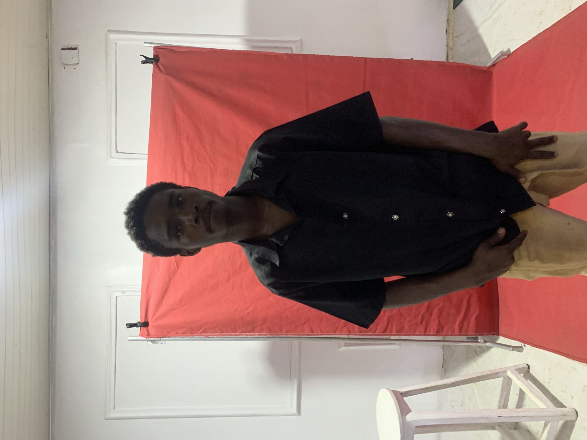 A medium shot of a dark-skinned person with short, curly hair wearing a black short-sleeved shirt and light brown pants stands in front of a red backdrop. The person is oriented diagonally in the frame with their head towards the left and their body angled towards the right. The background consists of white walls with what appears to be a white door frame behind the red backdrop. A small white stool is visible in the lower right corner. The photo appears to have been taken indoors. The location is not identifiable as Jos, Nigeria, from the image content alone.