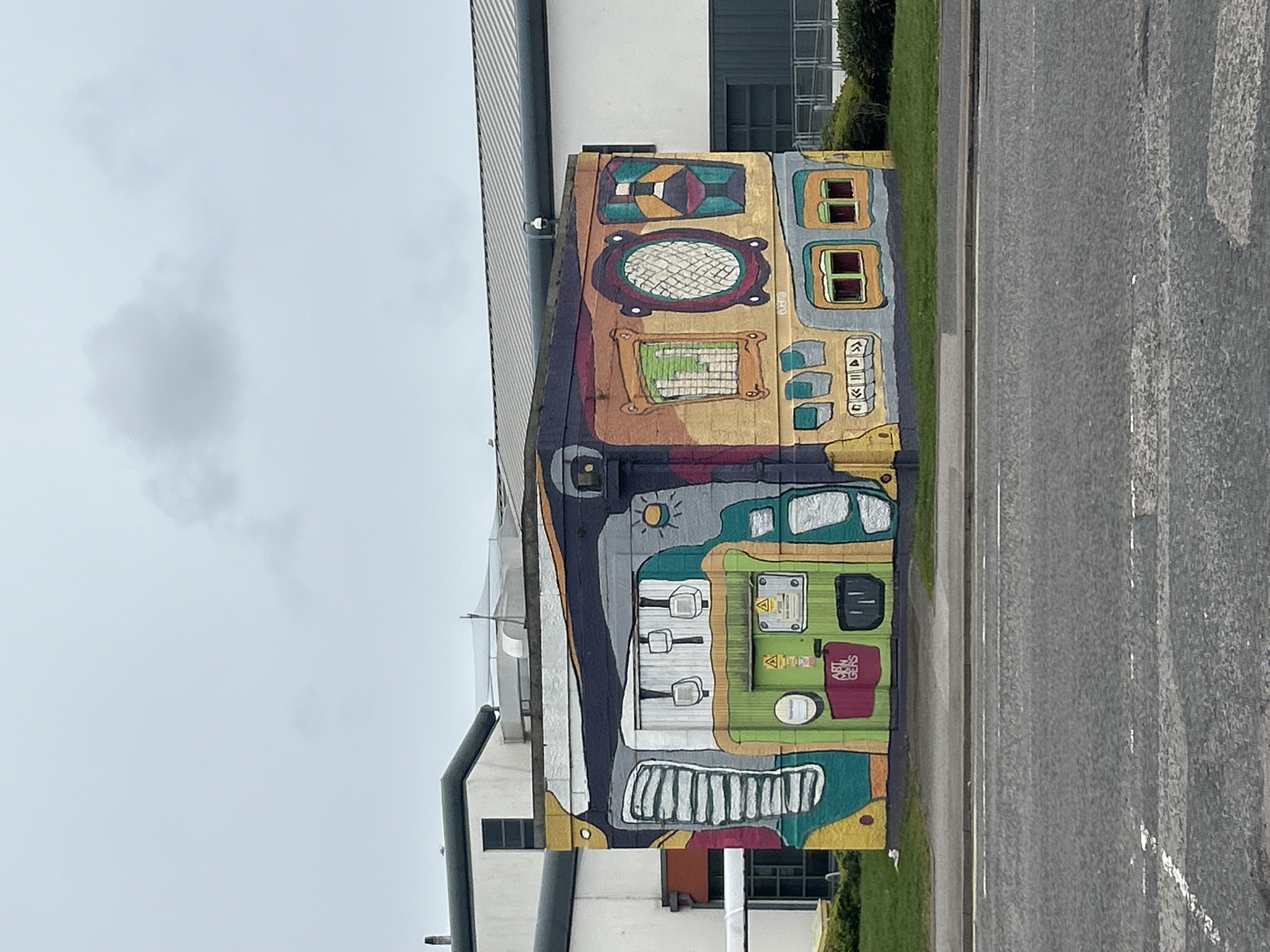 The image shows a utility box adorned with colorful, cartoon-style graffiti art. The artwork features multiple layers of abstract shapes and designs, mimicking the appearance of control panels, dials, and various technical elements. The box is situated on the side of a road, adjacent to a building with a corrugated metal exterior. The environment includes gray asphalt and green grass. This is located within Aberdeen City, United Kingdom. There are no people visible in the image.