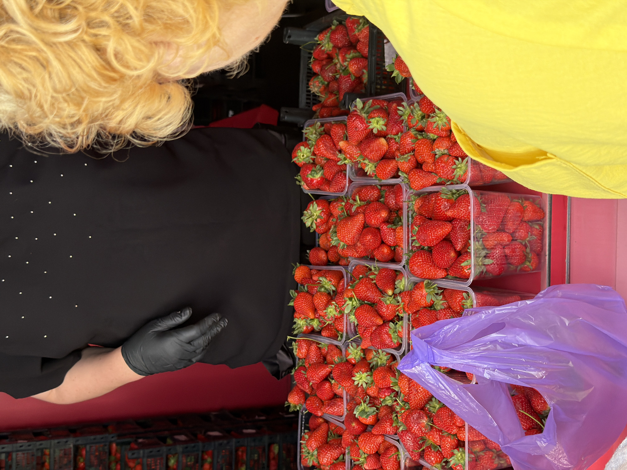 The image shows a market stall in Khimki, Russia, selling strawberries. There are several clear plastic containers filled with fresh, red strawberries. A person with blonde hair and wearing a black shirt with small, shiny embellishments and black gloves is visible to the left of the frame. Another person wearing a yellow shirt is partially visible to the right. A purple plastic bag is among the strawberries.