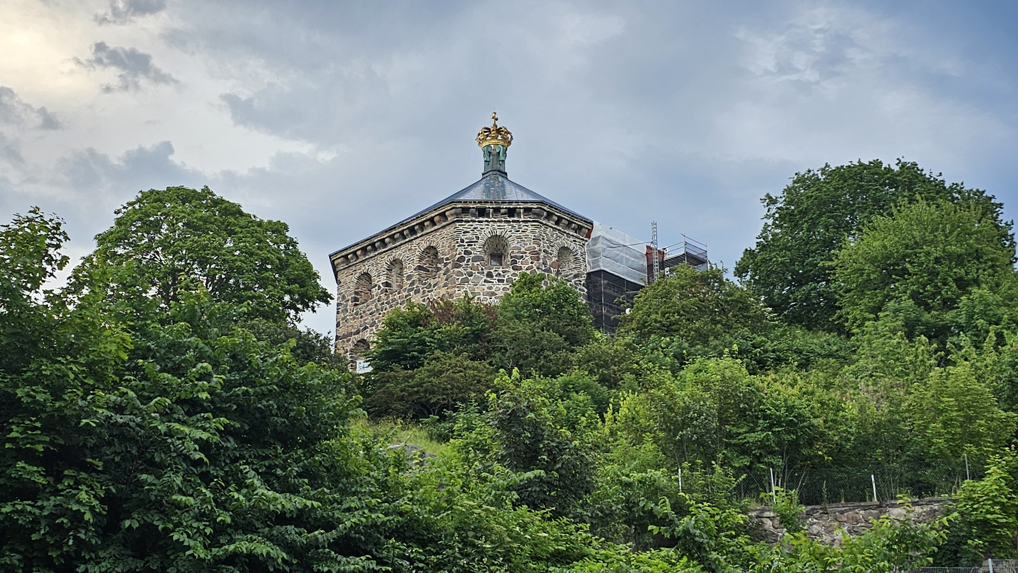The image depicts Skansen Kronan, a historic fortress located in the city of Gothenburg, Sweden. The fortress, constructed of stone, stands prominently on a hilltop, partially obscured by dense green foliage from surrounding trees and shrubs. The fortress features an octagonal structure with arched windows. A golden crown is visible atop its roof. Scaffolding and protective sheeting suggest ongoing construction or maintenance work on a section of the fortress. The sky above is overcast with scattered clouds, indicating a temperate weather condition. The foreground is filled with lush vegetation, a stone wall, and a wire fence. No people are visible in the scene.