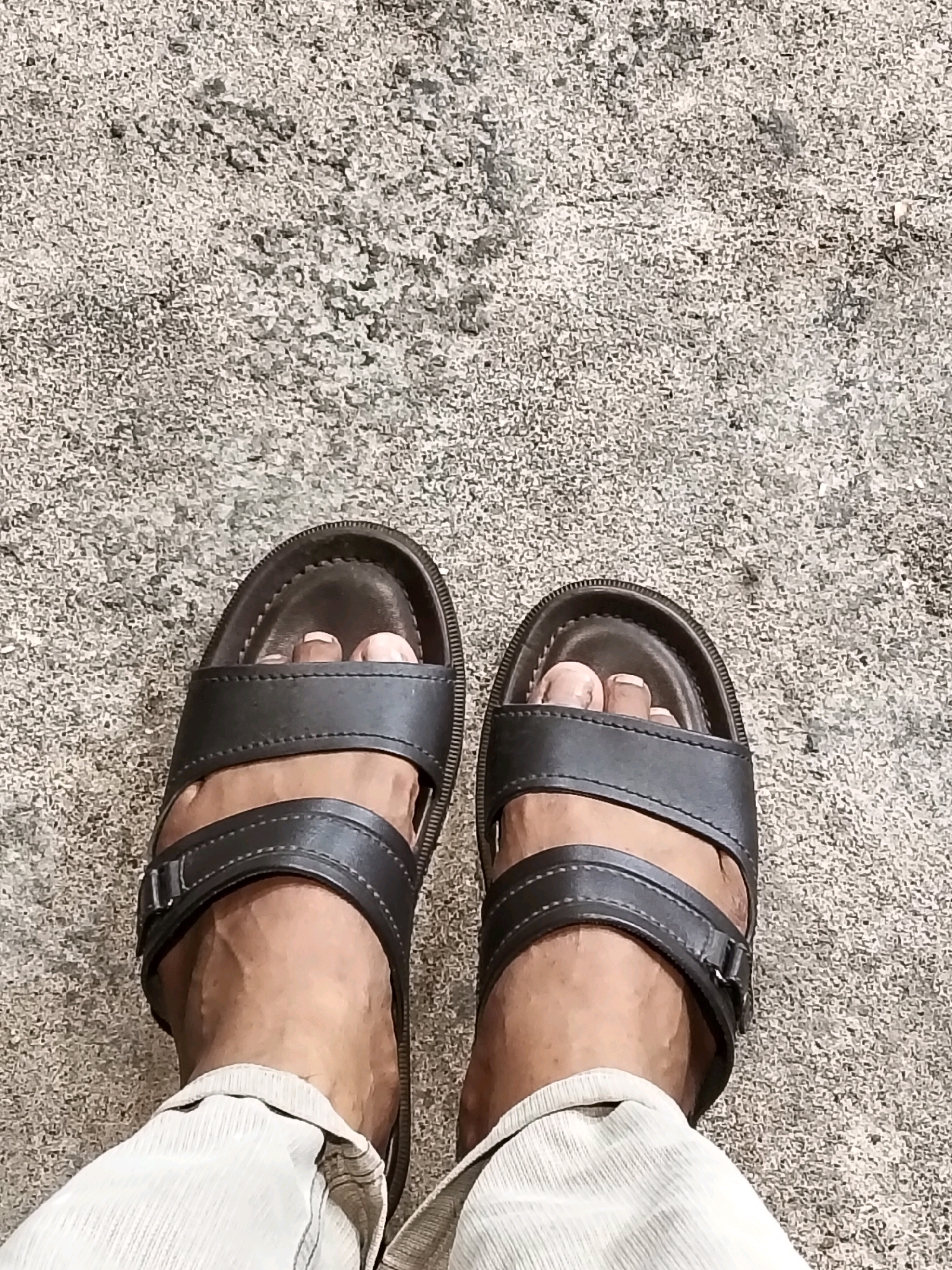 The image shows a close-up, overhead view of a person's feet wearing dark brown, two-strap sandals. The sandals are resting on a rough, grey textured surface, likely asphalt or concrete, suggestive of an outdoor urban environment in Arang Limbung, Indonesia. The person is wearing light-colored pants that partially obscure the lower legs. The lighting indicates a bright day, casting subtle shadows. The focus is on the footwear and the ground.