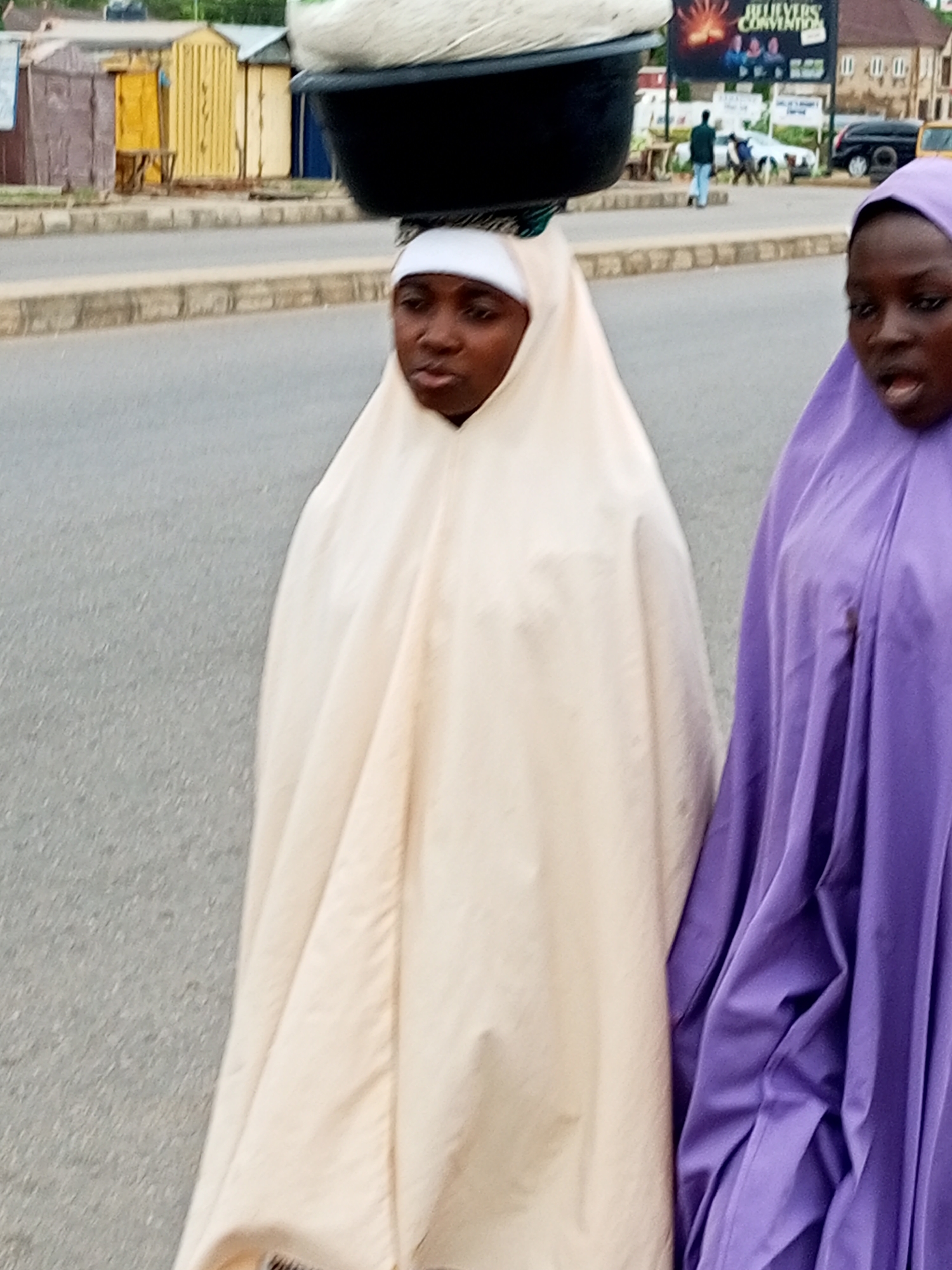 The image captures a street scene in Tumfure, Nigeria. Two women are the primary subjects. The woman on the left is wearing a cream-colored hijab and a head wrap, with a large black basin containing white material balanced on her head. She is looking forward with a neutral expression. The woman on the right is wearing a purple hijab, her mouth is open, and she appears to be speaking or calling out. They are walking on a paved road, with a sidewalk and some buildings visible in the background. The buildings appear to be small shops or stalls, painted in various colors including yellow, brown, and blue. A billboard with text and images is also visible in the background, as well as a black SUV and a yellow vehicle. The overall environment suggests an outdoor, possibly commercial area within the city.