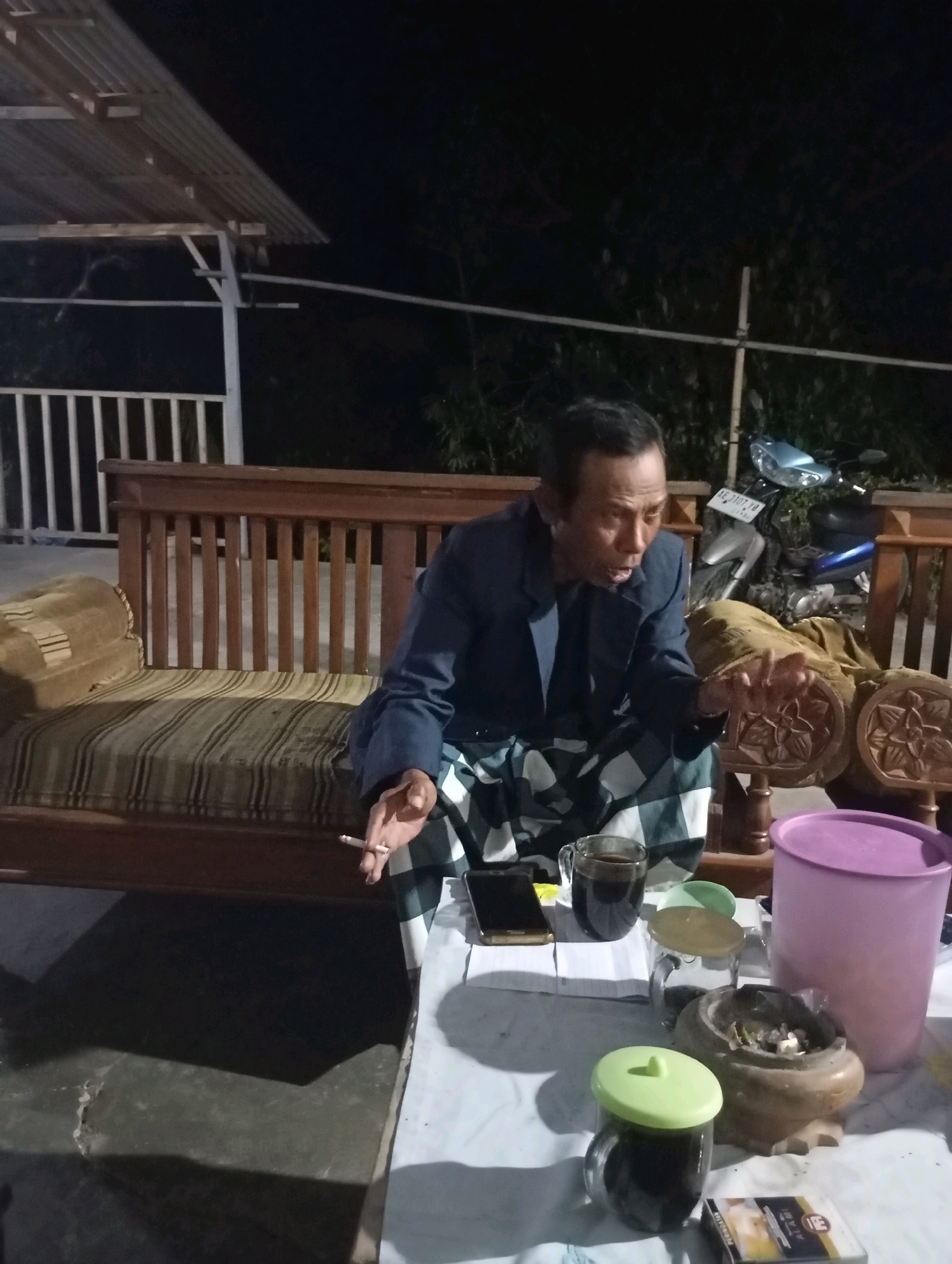 In Wonogondo, Indonesia, a man is seated on a wooden bench outdoors at night. He is wearing a dark suit jacket and a sarong. He is holding a lit cigarette in his right hand and appears to be speaking. In front of him, on a table, are a mug of dark liquid, a smartphone, a jar with a green lid, an ashtray with cigarette butts, and a pack of cigarettes. A large purple container is also visible on the table. The background shows a wooden structure and foliage. A motorcycle is parked behind the man.