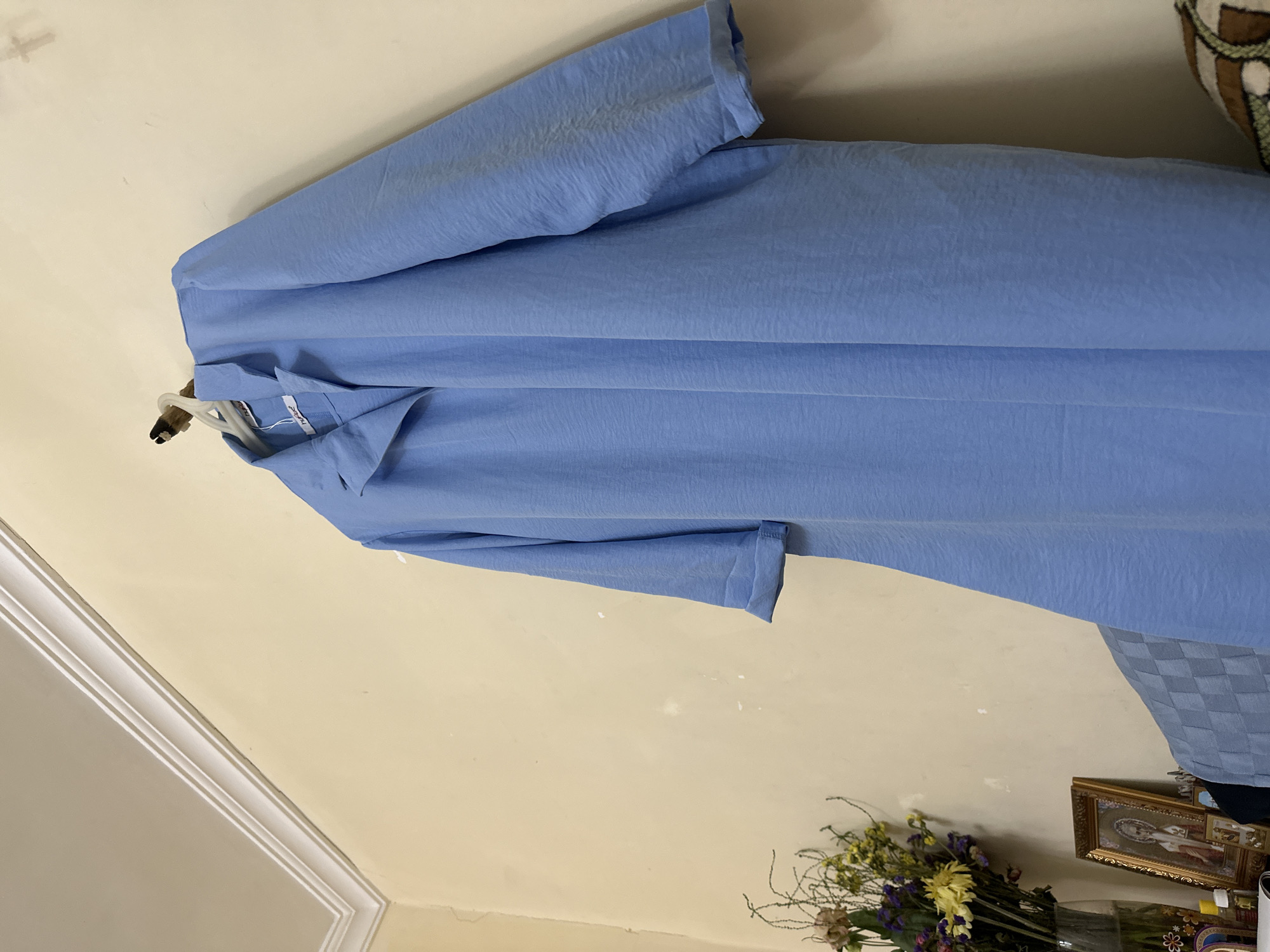 The image shows a light blue, long-sleeved robe or dress hanging on a hook against a cream-colored wall. The fabric of the garment has a textured, crinkled appearance. To the left of the garment, a vase of dried flowers and a framed religious icon are visible on a surface that appears to be a table or shelf. Above the garment, a white crown molding runs along the corner where the wall meets the ceiling. The scene appears to be indoors, likely in a residential setting in Yerevan, Armenia, based on the presence of the religious icon, which is common in Armenian homes. No people are present in the image.