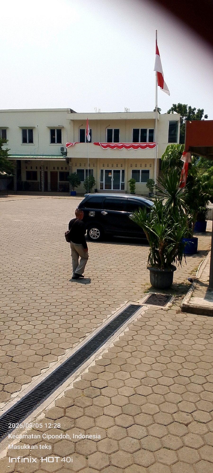 A person, identified as male based on clothing and build, walks across a paved courtyard in front of a light-colored, multi-story building. The building has several windows and is decorated with red and white bunting. An Indonesian flag flies from a flagpole on the roof. A black van is parked in the courtyard. The courtyard is paved with hexagonal stones, and a drainage grate runs along the ground. The scene is brightly lit by sunlight. The date stamp indicates the photo was taken on September 5, 2026, at 12:28 PM, in Cipondoh District, Indonesia.