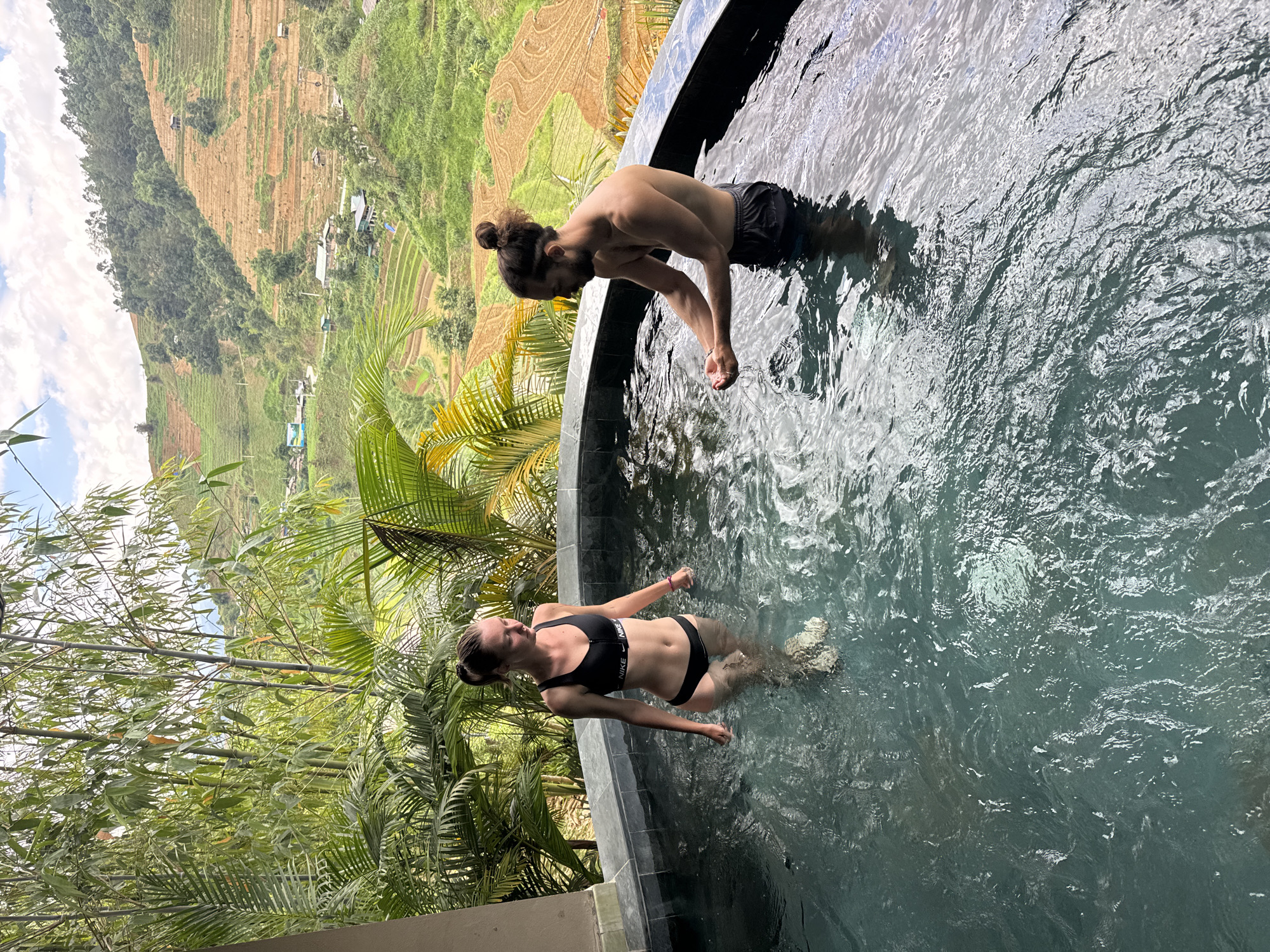 An outdoor, eye-level shot depicts a man and a woman in a dark-tiled circular hot tub or small pool, set against a backdrop of lush green foliage and terraced mountainsides in what is consistent with Mù Cang Chải, Vietnam. The woman, on the left, stands hip-deep in the water, facing right and looking down, wearing a black Nike sports bra bikini top and matching bottoms. A pink wristband is on her right wrist. The man, on the right, stands waist-deep, facing left and leaning forward, cupping water in his hands. He has dark hair tied in a top knot, a beard, and wears dark swim shorts. A light-colored bracelet is on his left wrist. The water is clear, showing ripples and reflections, and the individuals' legs are visible through it. Behind the pool, a dense array of green bamboo and large-leafed tropical plants frames the scene. In the distant background, steep hills are sculpted with numerous rice terraces, exhibiting a mix of reddish-brown soil and vibrant green vegetation, with scattered small dwellings and a winding road. The sky above is partly cloudy.
