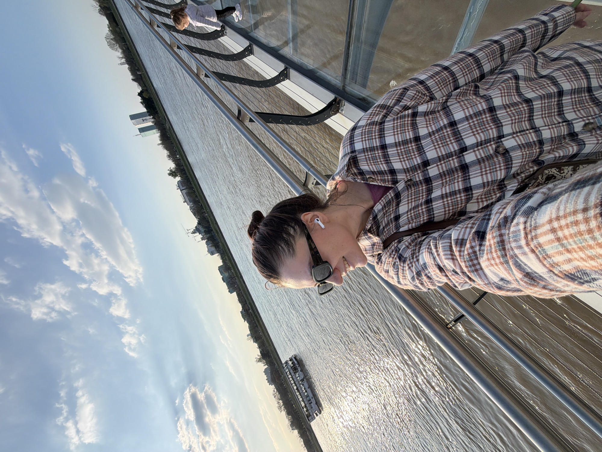 The image shows a woman taking a selfie on the glass floor of a pedestrian bridge over the Sava River in Belgrade, Serbia. A second person is visible in the background on the other side of the bridge. The Sava River and the city skyline are visible in the background, along with buildings, trees, and boats on the river. The sky is blue with clouds, and the sun is reflecting off the river.