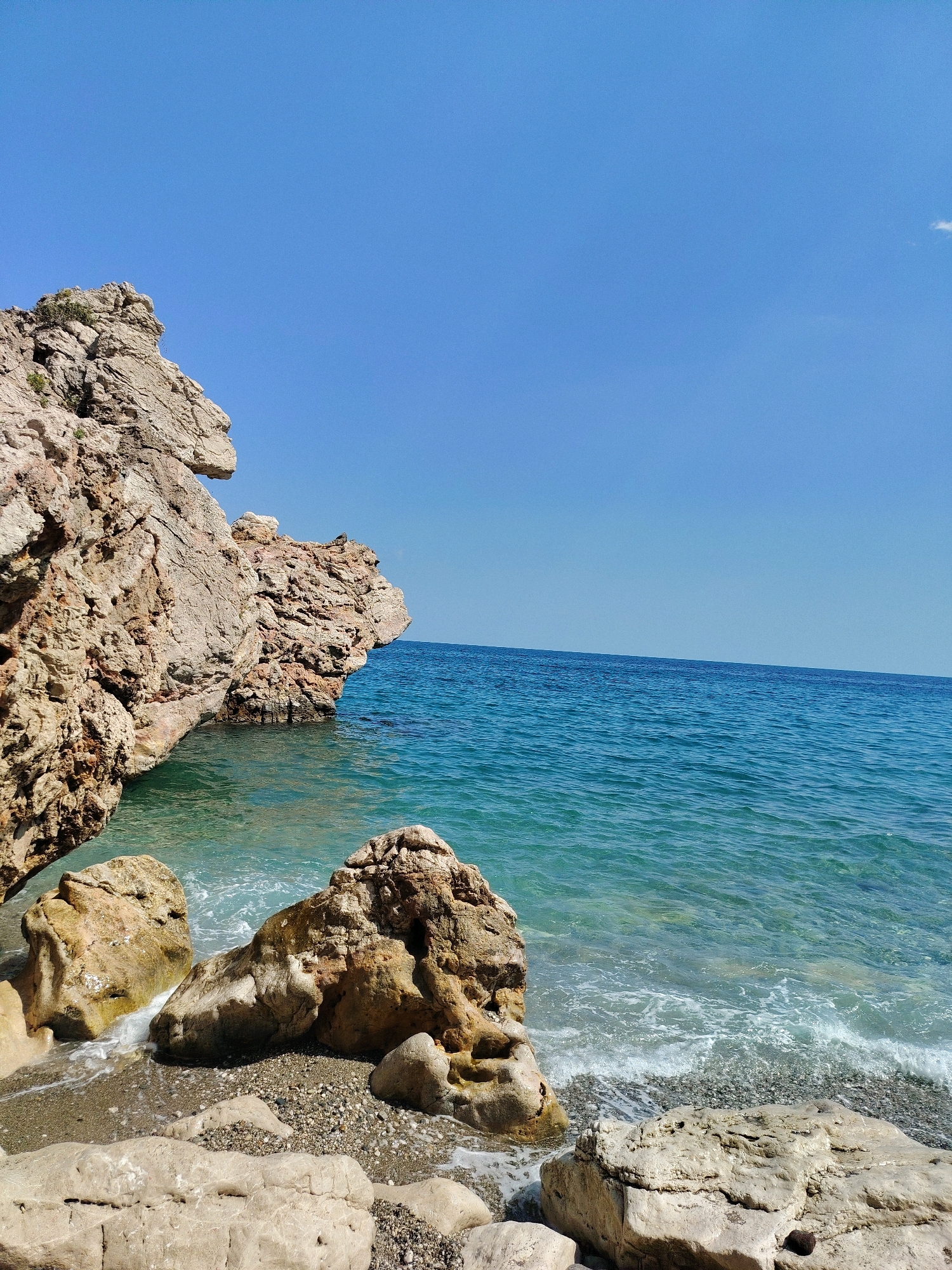 The image depicts a scenic view of a rocky coastline meeting the turquoise waters of the Mediterranean Sea. Large, weathered rock formations dominate the left side of the frame, casting shadows on the pebble-strewn shore below. The water is clear, with gentle waves lapping at the beach. In the distance, the horizon is a sharp line separating the sea from a vast, cloudless blue sky. The scene is likely located in or around Kemer, Turkey, known for its beautiful beaches and dramatic coastal landscapes. There are no people or any discernable actions or interactions present in the image. The environment is natural and serene, characterized by sunlight illuminating the rock textures and the shimmering surface of the sea.