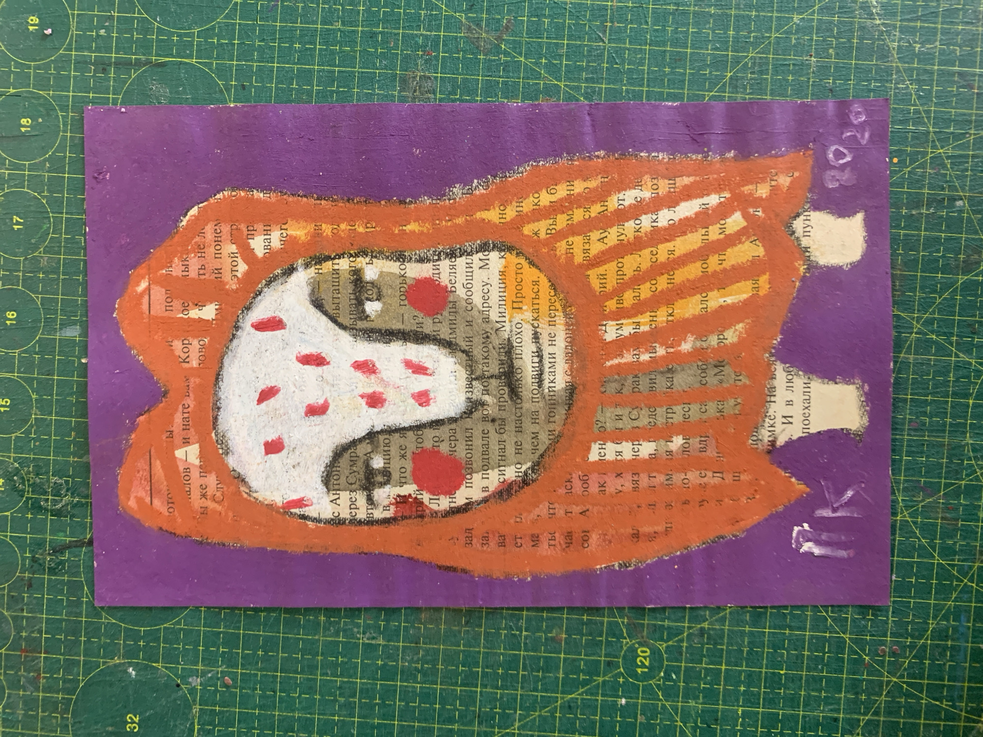 The image shows a small, vertical, mixed-media artwork on a purple background, placed on a green cutting mat with visible grid lines. The artwork depicts a stylized, owl-like creature with a white face adorned with red dots. The creature is outlined in orange and appears to be wearing a large, hooded orange garment. The background of the creature is composed of torn pieces of text from a book, visible through the painted elements. Below the creature's feet, there is a white painted area with the initials "RK" and the year "2020" also painted in white.
The cutting mat is a standard craft tool, often used for precise cutting of materials. The grid lines on the mat help with measurements and alignment. The overall scene is that of an artist's workspace or a studio.
The question asks for the location in the city of Sergeevo, Russia. However, there is no visual information within the image or its accompanying crops that indicates the location is Sergeevo, Russia. Therefore, this detail cannot be provided based on the visual evidence.