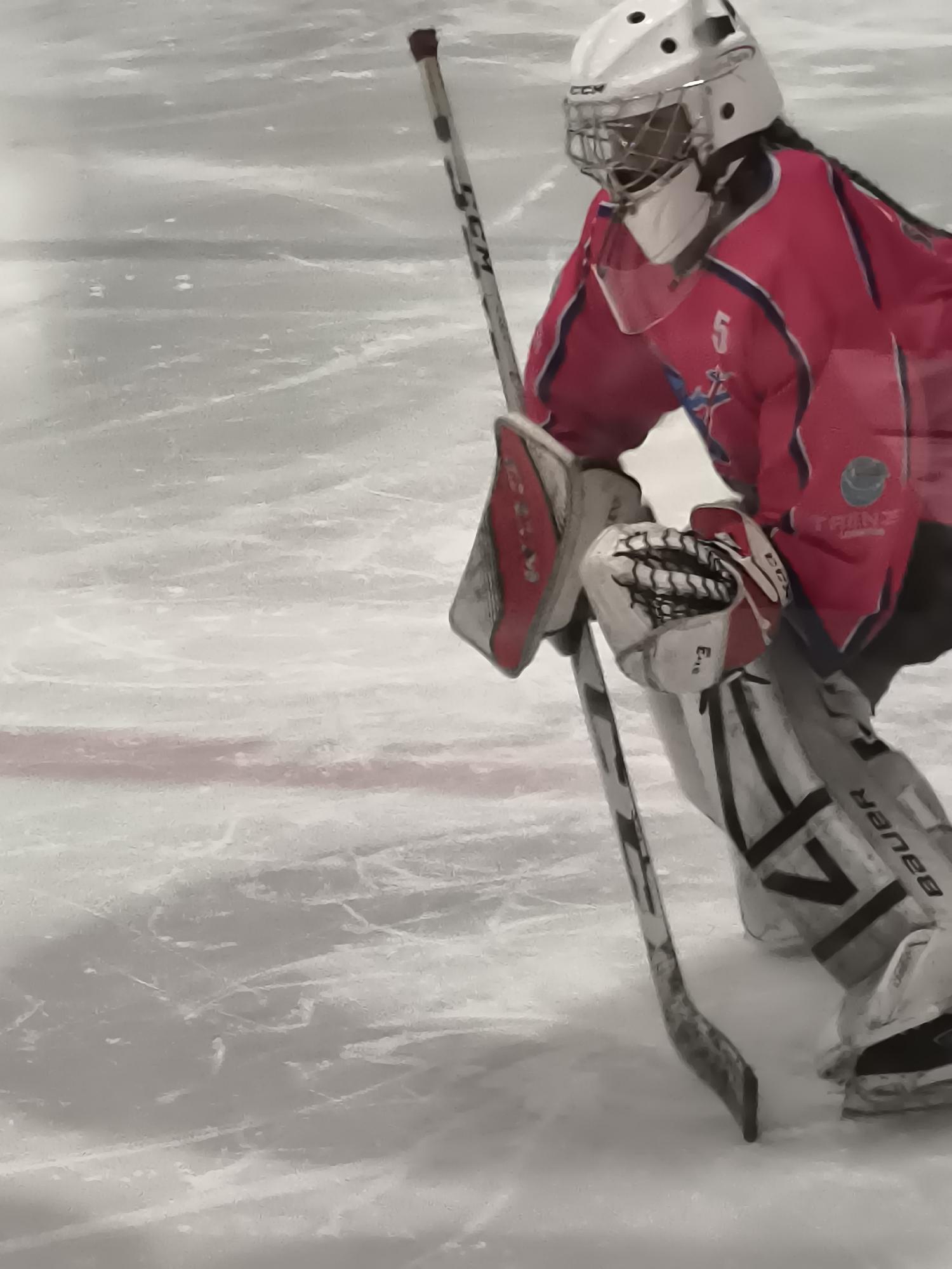 A hockey goalie, wearing a pink jersey, white helmet with a cage, and protective gear, stands on an ice rink. The goalie holds a hockey stick in their right hand, with the blade resting on the ice. The goalie's left hand is gloved, with the glove positioned in front of their body. The ice rink has visible skate marks and a pink line is visible near the bottom left of the frame. The background is out of focus, showing the icy surface. There is no discernible information to indicate the location in the city of Klaipėda, Lithuania.