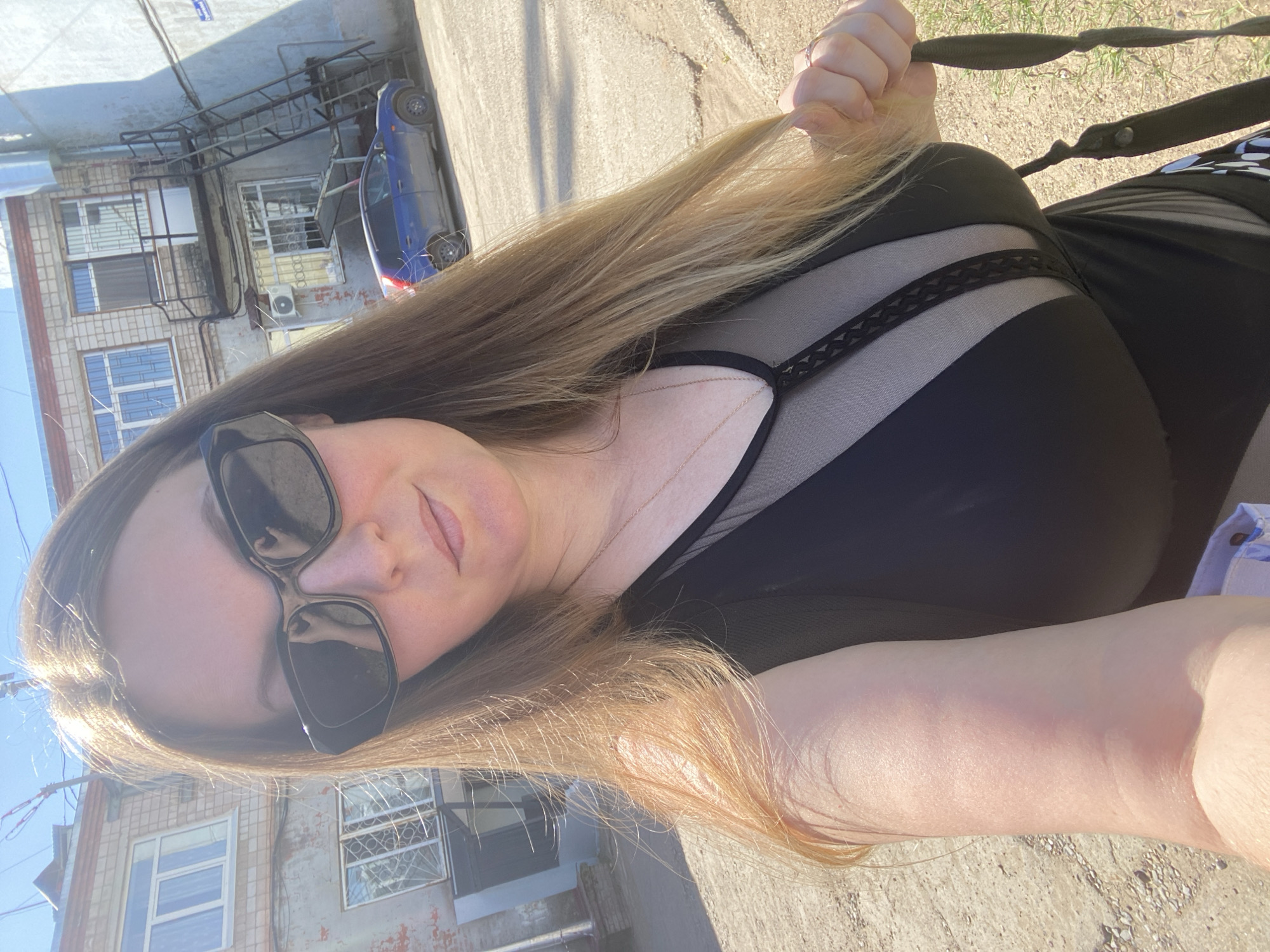 The image shows a woman wearing a black dress with sheer panels, sunglasses, and a gold necklace taking a selfie outdoors. She is holding a strap or handle in her right hand. The background includes a building with windows, an external fire escape, and a parked blue car. The location is Noviy, Russia, judging by the architecture and the vehicle registration plate visible in the full resolution image. The setting appears to be a paved area next to a building. The woman's hair is blonde and falls over her shoulder. The sun appears to be overhead, casting shadows.