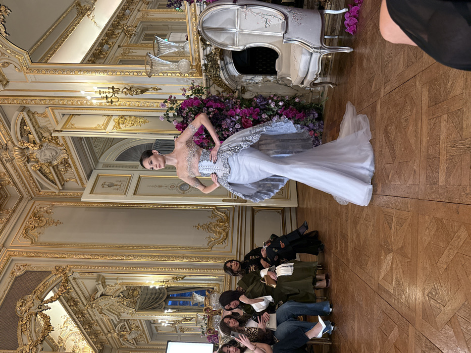 A model in a striking silver gown poses indoors amidst ornate gold décor and seated onlookers. The dress features a shimmering bodice and a flowing skirt, complemented by elaborate detailing. A floral display adorns the background, accentuating the luxurious setting. The audience is seated below the model with their eyes looking at the model. The room's design includes mirrors, detailed molding, and parquet flooring.