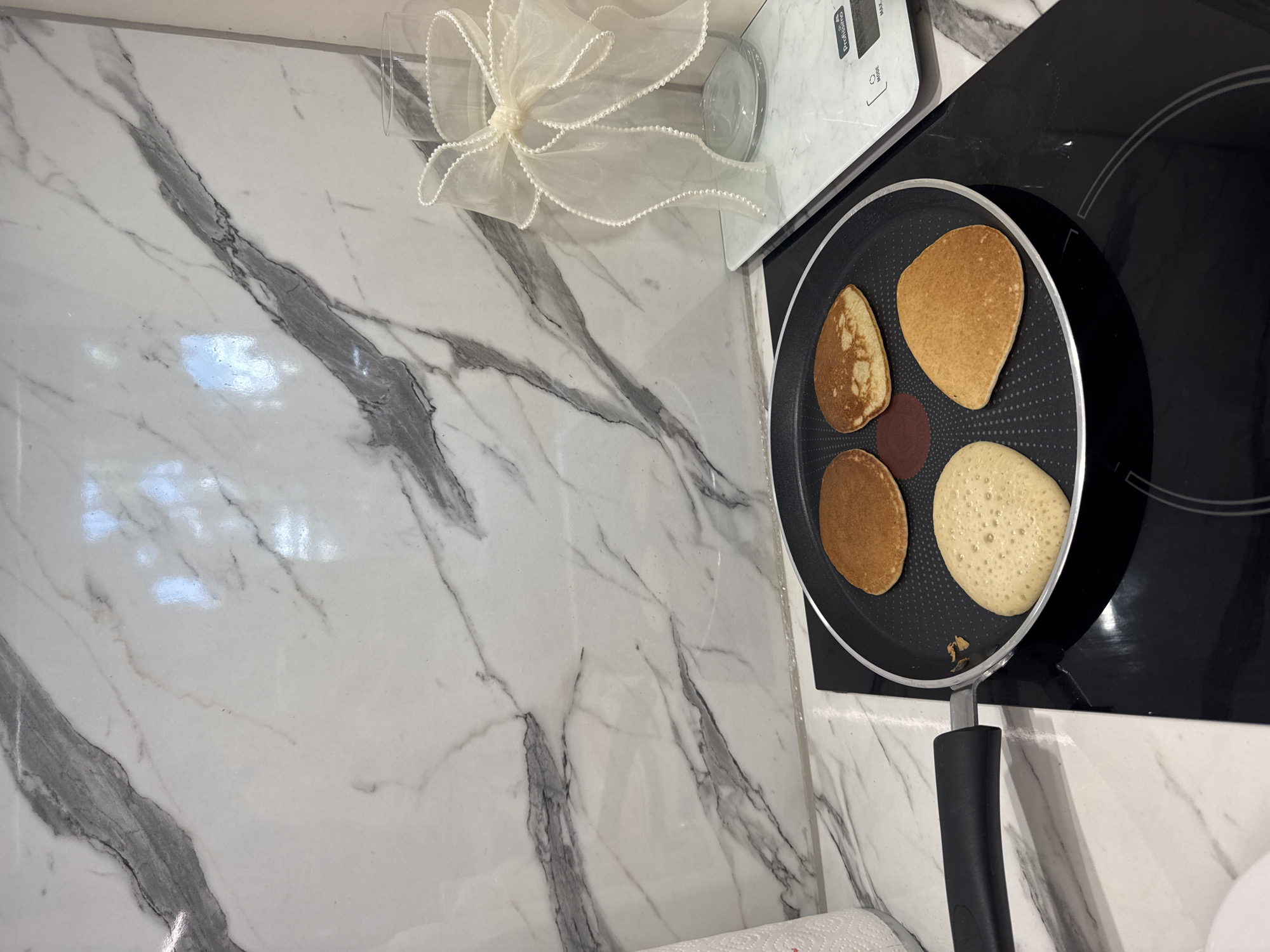 In an indoor kitchen setting, likely a modern apartment, four pancakes are cooking in a black non-stick frying pan. The pan rests on a black induction stovetop, which is part of a white countertop. The background features a marble-patterned backsplash. To the right of the stovetop, a white digital scale displays the brand "Profissimo." A clear glass vase with a decorative sheer ribbon tied around its neck is placed on the scale. To the left of the pan, a roll of paper towels is partially visible. The lighting suggests an interior setting, possibly daylight from a nearby window, as evidenced by reflections on the marble. There are no people present in the scene. The image is a close-up shot, focusing on the cooking process and the immediate kitchen environment. The location is not specified to be in Sofia, Bulgaria.