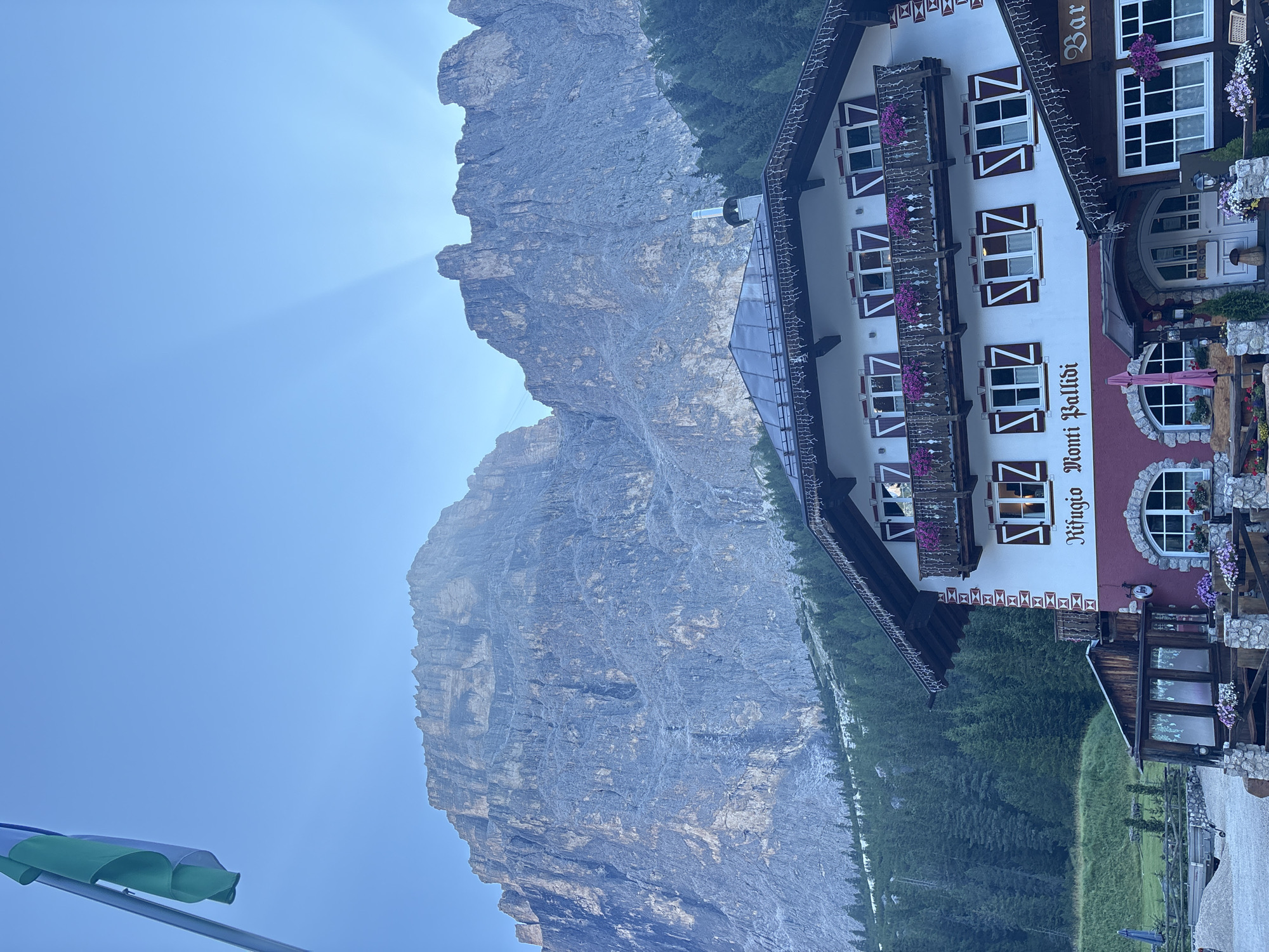 The image shows a multi-story building with a traditional Alpine architectural style. The building has a white facade with red lower levels and dark brown trim. It features multiple windows with red shutters and balconies adorned with flower boxes filled with purple flowers. Text on the building reads "Rifugio Monti Pallidi" and "Bar." A flag, appearing to be green and blue, is visible in the upper left corner, attached to a flagpole.
In the background, a massive, rugged mountain range dominates the landscape. The peaks are steep and rocky, with visible textures and striations. Sunlight is breaking through the clouds, creating dramatic light rays illuminating the mountain face. Pine trees cover the lower slopes of the mountains, creating a dense forest.
The foreground shows a gravel-covered area, suggesting a pathway or parking area. A wooden fence with stone pillars encloses part of the area, and a trailer is visible on the left. Various potted plants and flowers are placed around the entrance of the building, adding to its charm. The scene appears to be set in a mountainous region during daylight. The building is identified as Rifugio Monti Pallidi in Canazei, Italy, which is a mountain hut or lodge.