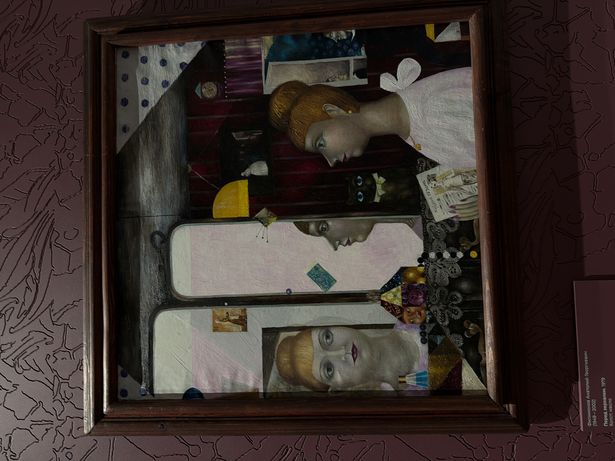 The image shows an oil painting in a brown wooden frame, hanging on a wall with a patterned aubergine-colored wallpaper. The painting depicts three reflections of a woman in a mirror. The first woman, at the top of the frame, is looking down with her red hair in a bun. She is wearing a white garment with a bow. Near her is a black cat with blue eyes wearing a white ribbon. Below her is a mirror image of the same woman with a card near her reflection. The third woman is looking up with a direct gaze. The label next to the painting identifies it as "Before the Mirror, 1979" by Anatoly Georgievich Filimonov (1949-2002). The location of the image is Perm, Russia.
