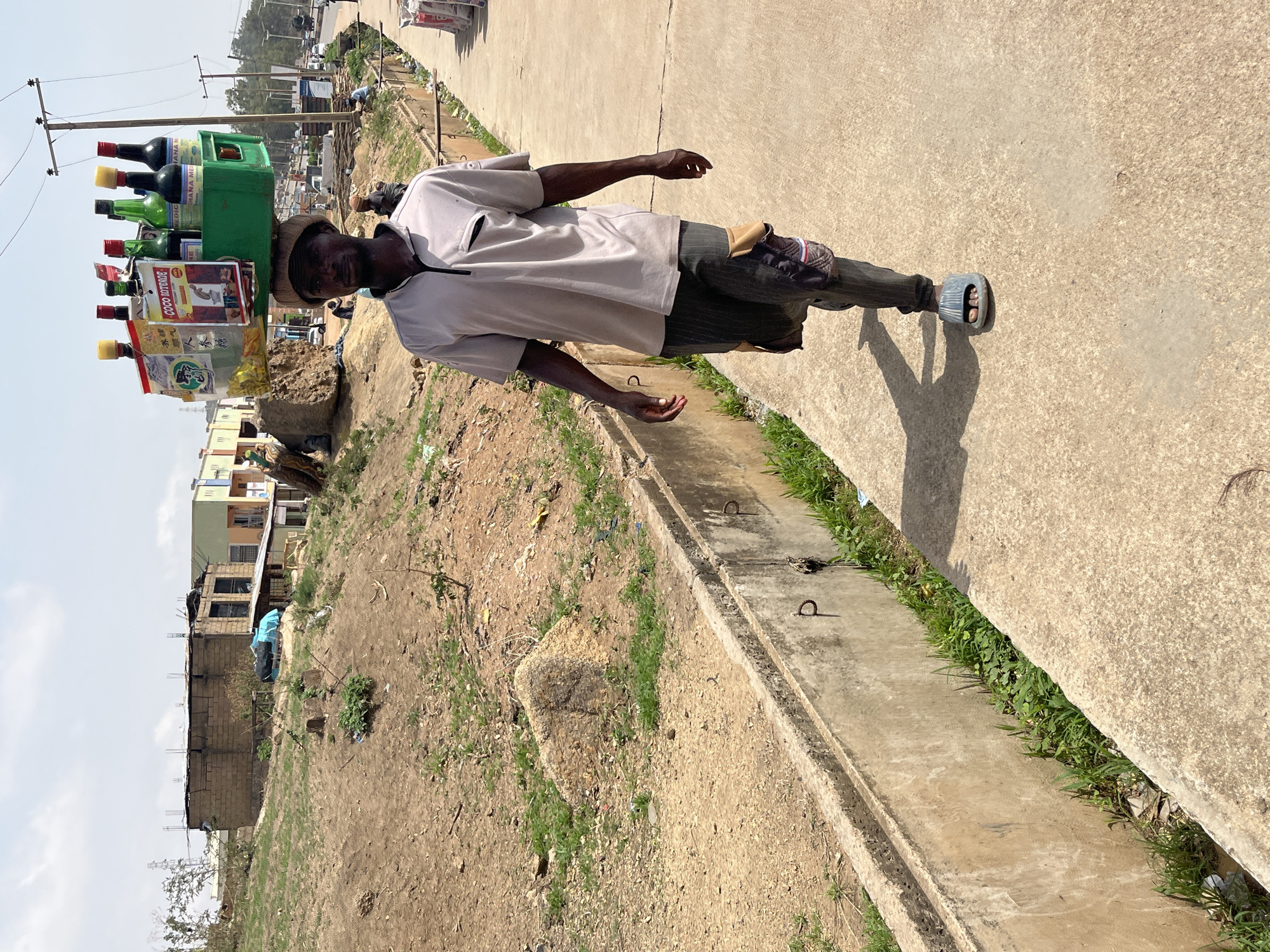 In Gura Top, Nigeria, a man stands on a concrete walkway beside a drainage ditch. He carries a green crate on his head, filled with bottles and small packages. He wears a light-colored shirt, dark pants, and sandals. The background shows a mix of buildings and open, grassy areas.