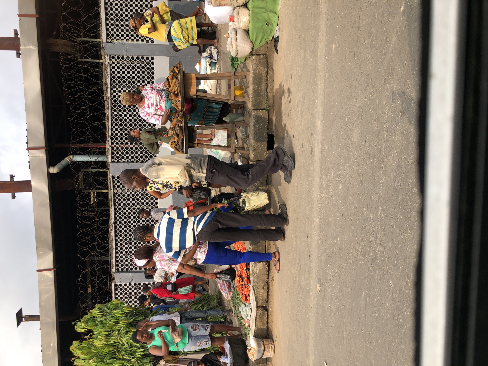 This image depicts a street market scene in Lagos, Nigeria, likely in a bustling area with a significant amount of pedestrian and commercial activity. The foreground shows a paved road with some debris and a distinct white line near the bottom edge, suggesting a vehicle or camera is positioned close to the ground.
In the mid-ground, a group of people are gathered around stalls and displays of goods. The atmosphere appears lively, with individuals interacting and engaged in commerce. Several vendors are visible, some behind tables laden with produce and other items. One woman in the center is wearing a brightly colored, patterned dress and appears to be a vendor selling what looks like dried fish or meat from a metal tray. Another woman in a pink and white patterned shirt and blue leggings is standing near her. To the left, a woman in a green tank top and ripped denim shorts is interacting with someone off-camera.
Various goods are on display, including piles of red tomatoes, leafy greens, and sacks of what could be grains or other foodstuffs. The stalls are set up on the sidewalk or raised platform adjacent to the road. Behind the market stalls, a building with a distinctive grey facade featuring circular cutouts is visible. Above this building, a dark fence with coiled barbed wire runs along the top, suggesting a security measure or boundary.
The overall environment suggests a typical urban street market in Lagos, characterized by informal vending, a mix of goods, and a dense population of people. The lighting indicates it is daytime, possibly late afternoon given the warm tones and shadows. The presence of greenery from a large bush or tree on the far left adds a touch of nature to the otherwise urban setting.